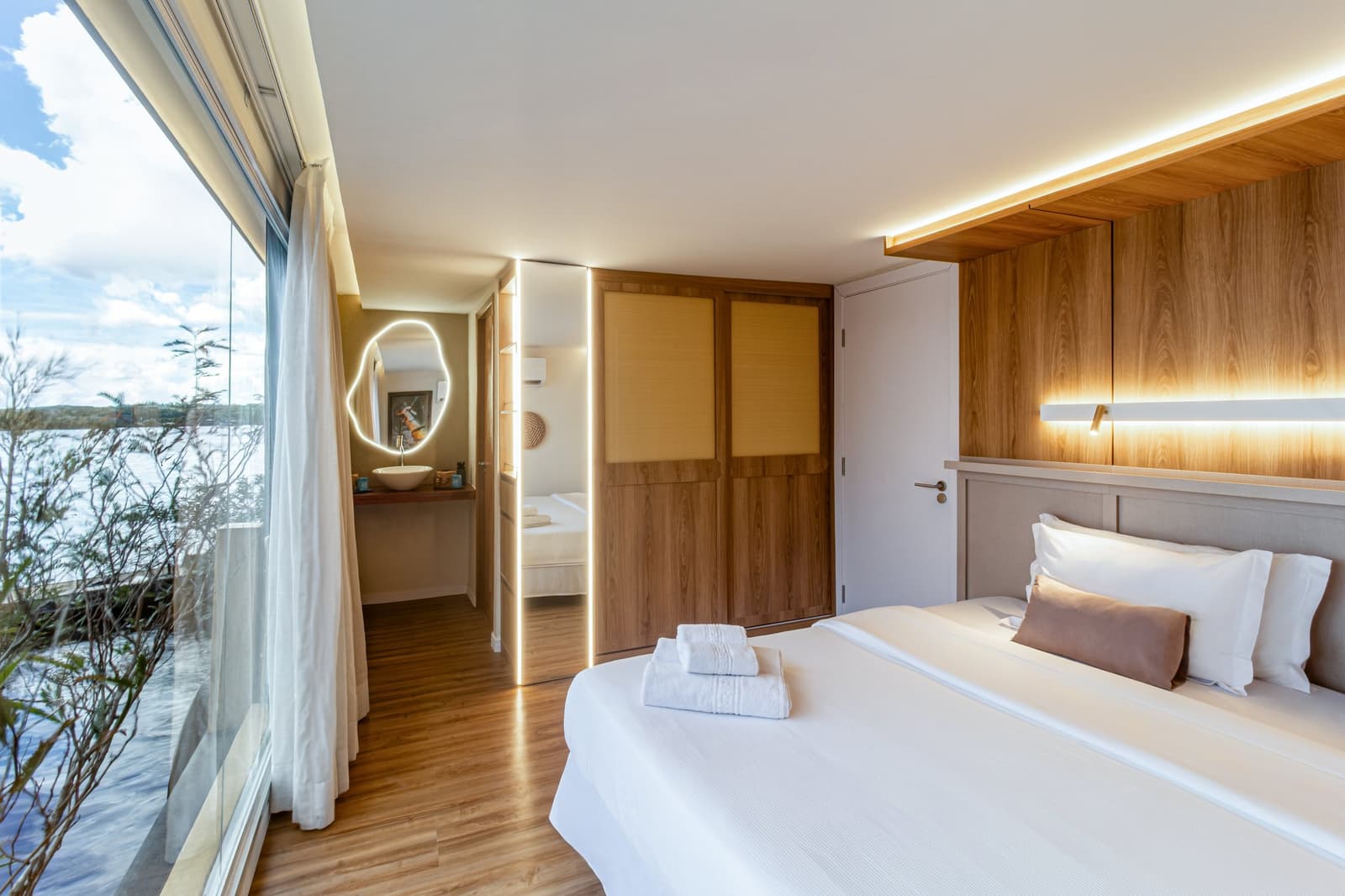 Your room, the river, and nothing in between. Photo: STUDIO CREATE Bright en-suite cabin on a luxury Amazon cruise in Brazil, featuring a king bed with white linens, warm wood-panelled headboard with LED lighting, floor-to-ceiling windows overlooking a wide river, and an open-plan area with an illuminated vanity mirror