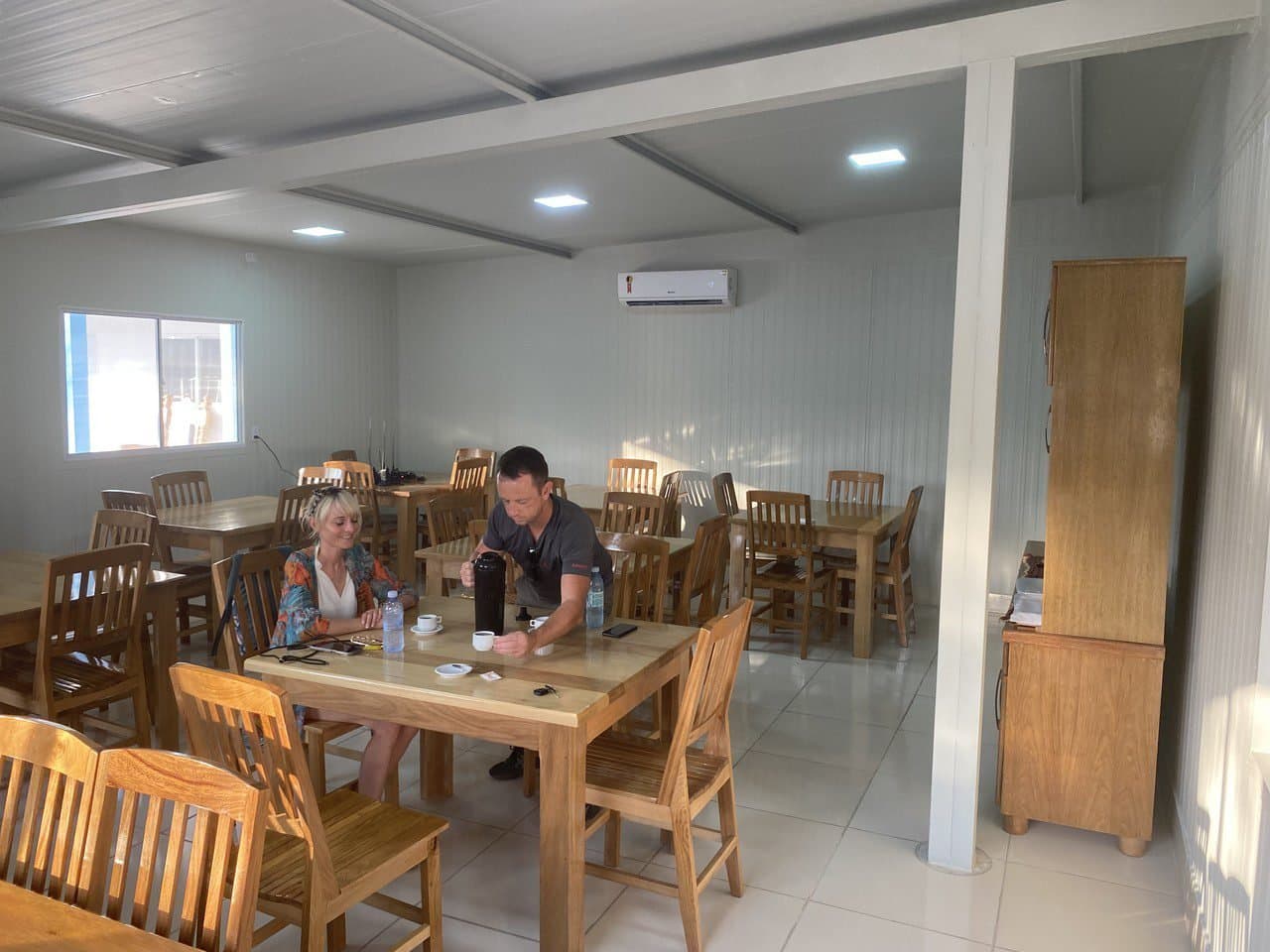 Good coffee and a quiet room before the wetlands take over. Two travellers sharing coffee at a wooden table inside a clean, air-conditioned dining room at a lodge on an amazon and pantanal tour, with rows of empty tables and chairs filling the bright space