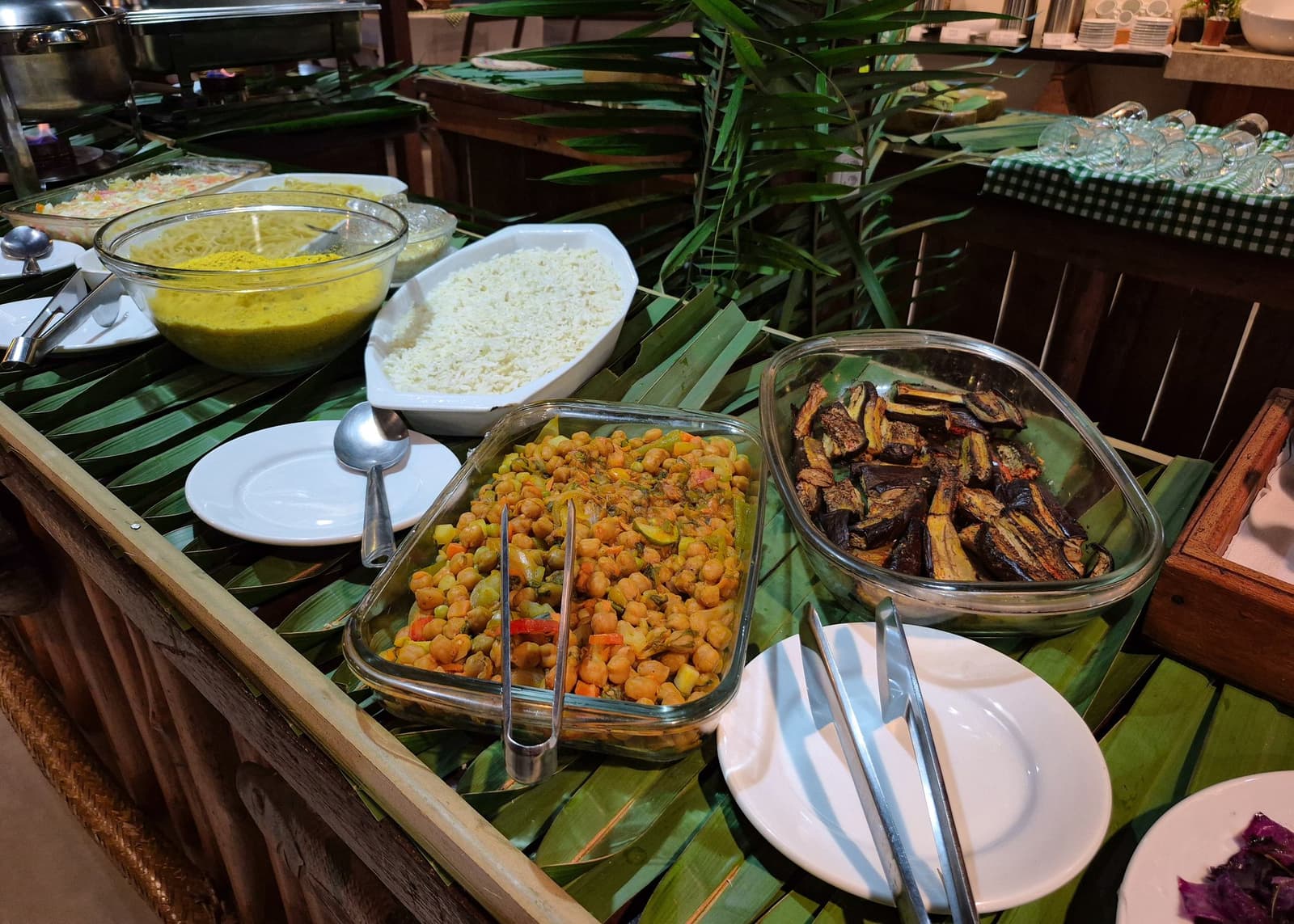 Honest, generous food, the kind that actually refuels you for the next safari. A rustic buffet table lined with fresh banana leaves holds glass dishes of spiced chickpeas, roasted eggplant, white rice, and a yellow sauce, set up at a lodge on an amazon and pantanal tour
