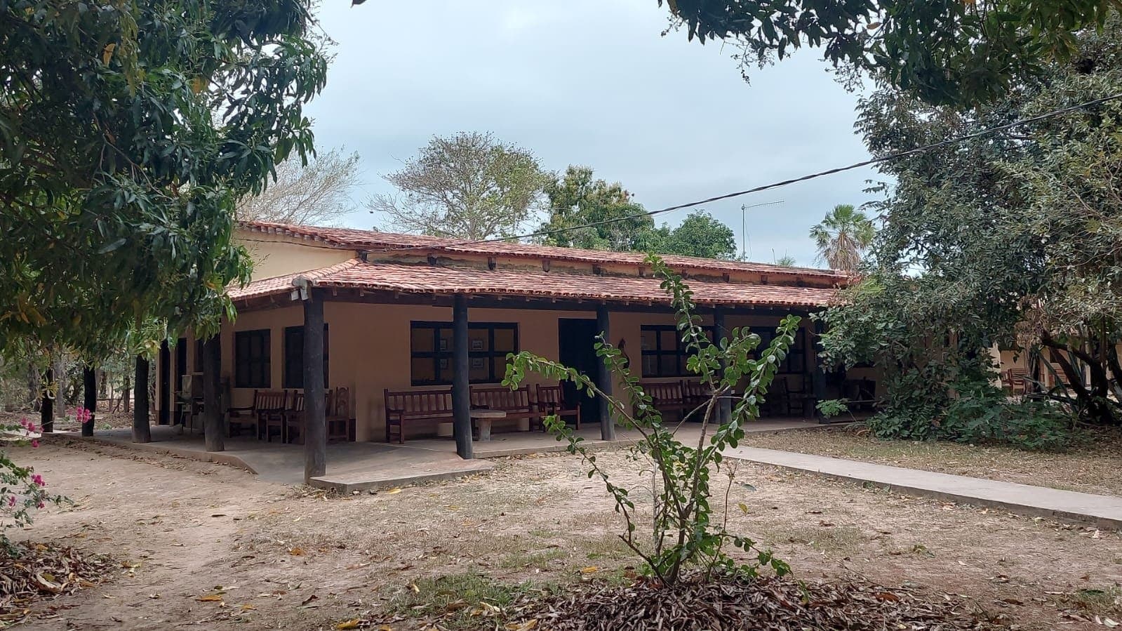 Bench, shade, and nothing urgent on the agenda, the Pantanal pace starts here. A low-slung ranch-style lodge with terracotta roof tiles and a shaded veranda lined with wooden benches, surrounded by dry-season trees on an amazon and pantanal tour stopover