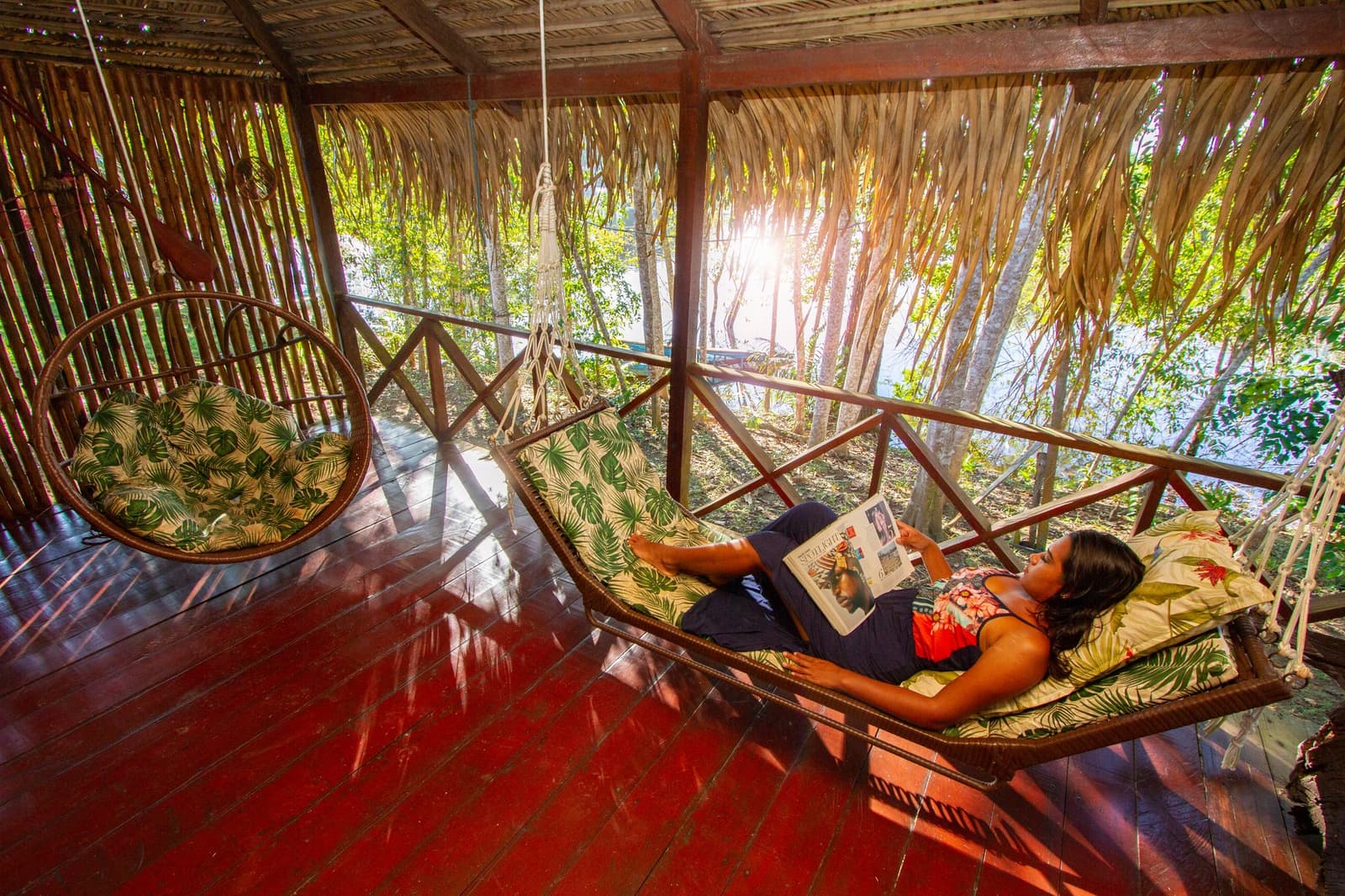 Some afternoons, the best thing to do is absolutely nothing. Photo: David Nascimento @ A woman lies on a cushioned hanging hammock bed reading a magazine on the open-air thatched terrace of an amazon jungle lodge, surrounded by lush forest and warm afternoon light