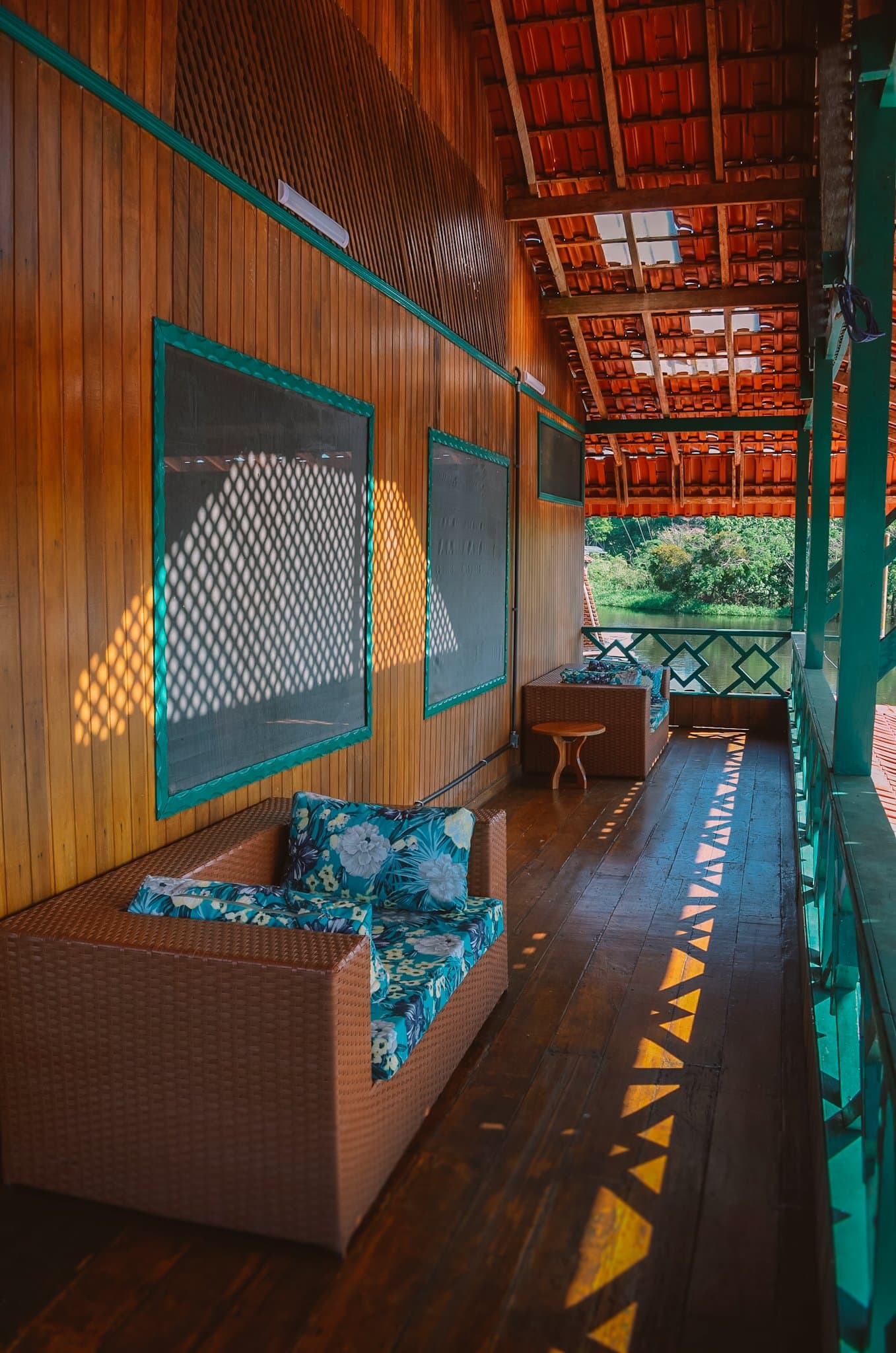 Afternoon light through the screens, the river just a few steps away. Covered wooden veranda of an amazon rainforest ecolodge trip in brazil accommodation, with wicker seating, floral cushions, screened windows casting lattice shadows, and a view of green riverbank vegetation beyond the railing