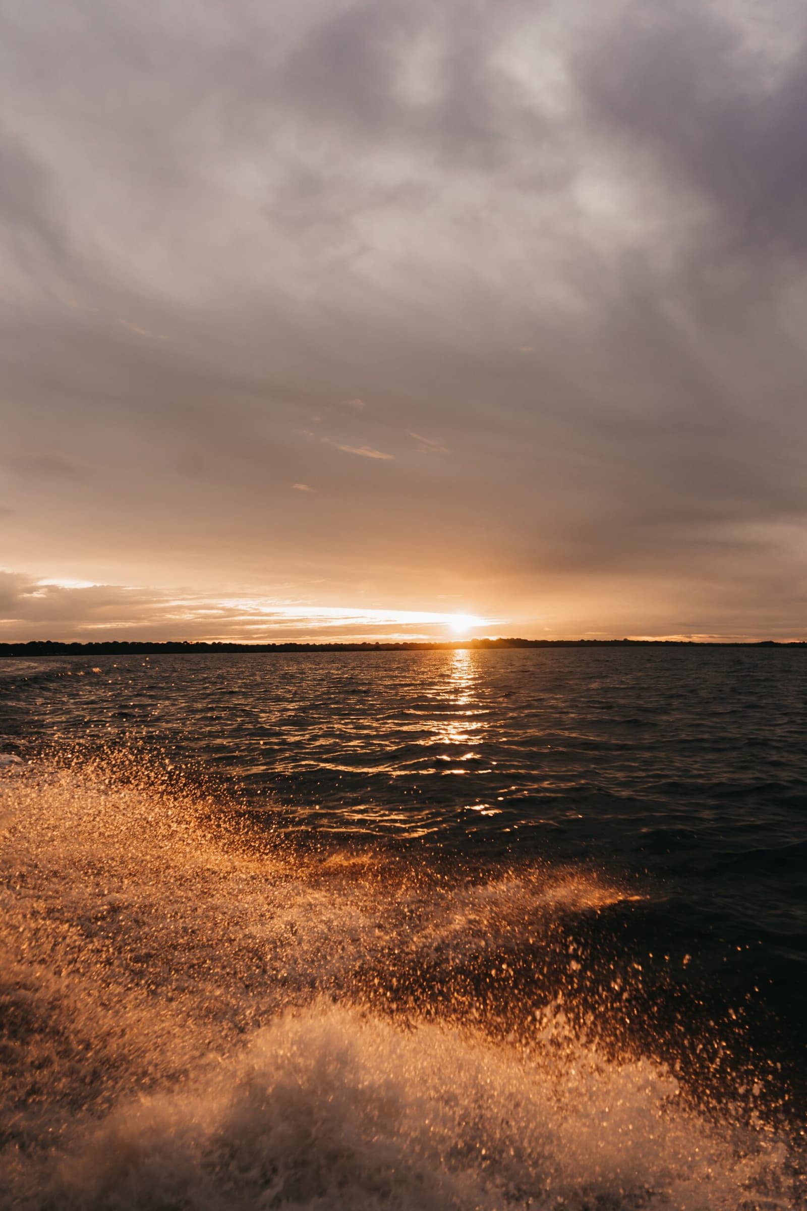 Golden hour hits differently when the river is throwing it back at you. Boat wake spray lit gold by a sunset over a wide river, with a distant treeline silhouetted beneath a dramatic cloudy sky on a luxury Amazon cruise in Brazil