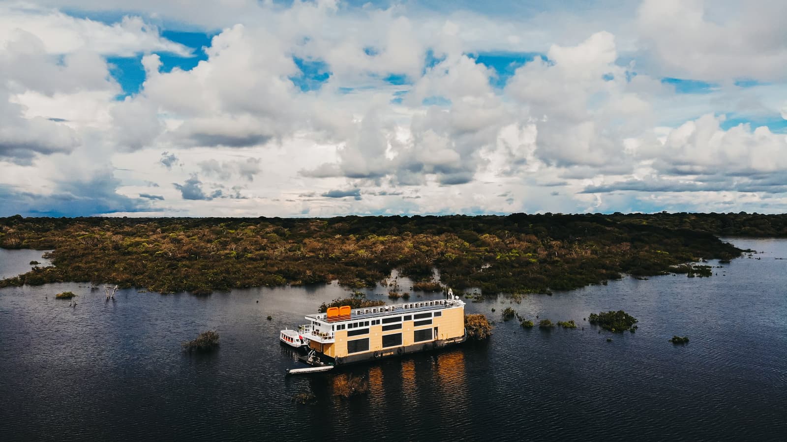 Your floating base, small against the forest, and that is exactly the point. Aerial view of a multi-deck luxury amazon cruise in Brazil moored on dark blackwater channels surrounded by flooded forest islands under a dramatic cloudy sky