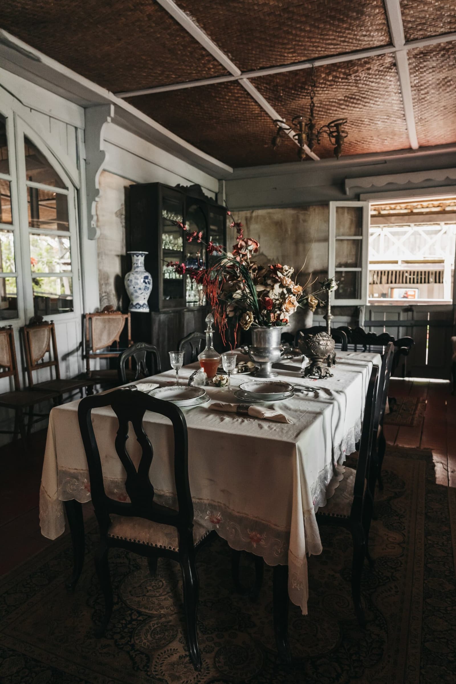 Dinner here comes with a century of rubber-boom stories baked into the walls. Historic dining room on a luxury Amazon cruise in Brazil, with a white linen-draped table set with silver and crystal, dark wood chairs, a blue-and-white porcelain vase, dried floral centrepiece, and a woven-panel ceiling under soft natural light