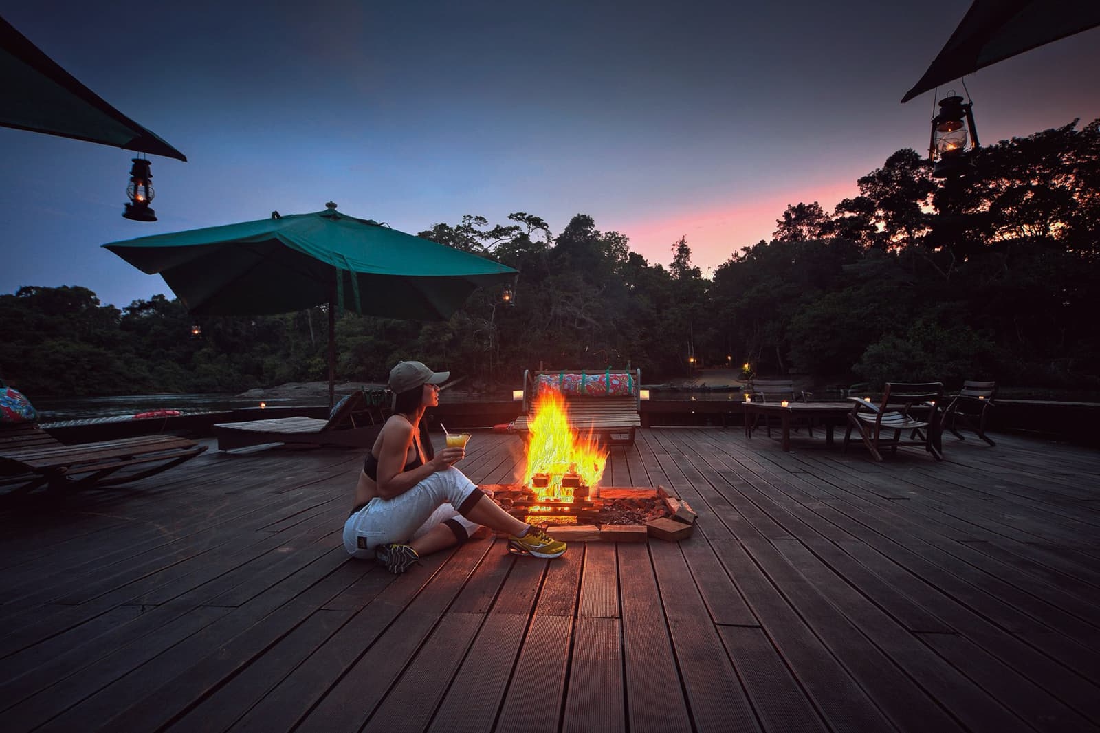 Drink in hand, fire going, forest right there. Some evenings at the lodge earn their reputation. Photo: Samuel Melim A woman sits cross-legged on a wide wooden deck beside an open fire pit during dusk, holding a drink, with hanging lanterns, a teal parasol, and dense forest silhouetted against a pink and blue twilight sky — an evening moment on a luxury Pantanal and Amazon trip