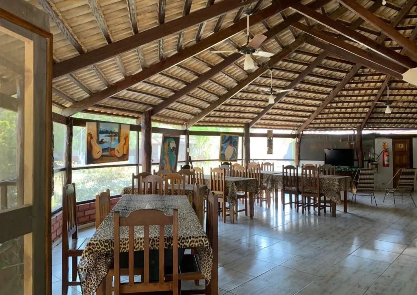 Lunch tastes better with jungle views on three sides. Open-air dining room at a Pantanal lodge on an amazon and pantanal tour, with wooden beam ceiling, ceiling fans, leopard-print tablecloths, and wildlife-themed paintings along the screened walls