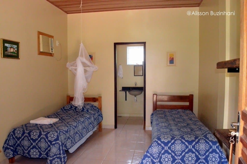 Clean, cool, and ready, the mosquito net is just there for good measure. Photo: Alisson Buzinhani Twin-bed room at a Pantanal lodge on an amazon and pantanal tour, with matching blue patterned bedspreads, a hanging mosquito net, air conditioning unit, and an open en-suite bathroom visible in the background