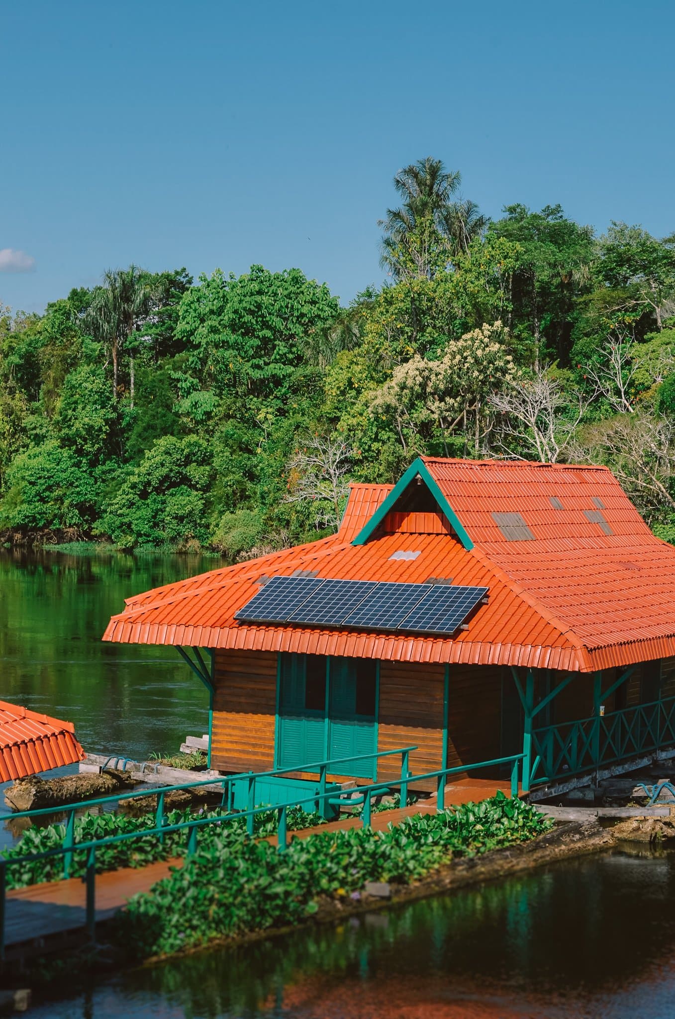 Off the grid, on the water, inside the forest. Not bad for a base camp. Solar-panelled floating wooden cabin with an orange tiled roof on a dark river, surrounded by dense tropical forest canopy — part of an amazon rainforest ecolodge trip in brazil at the Uakari floating lodge
