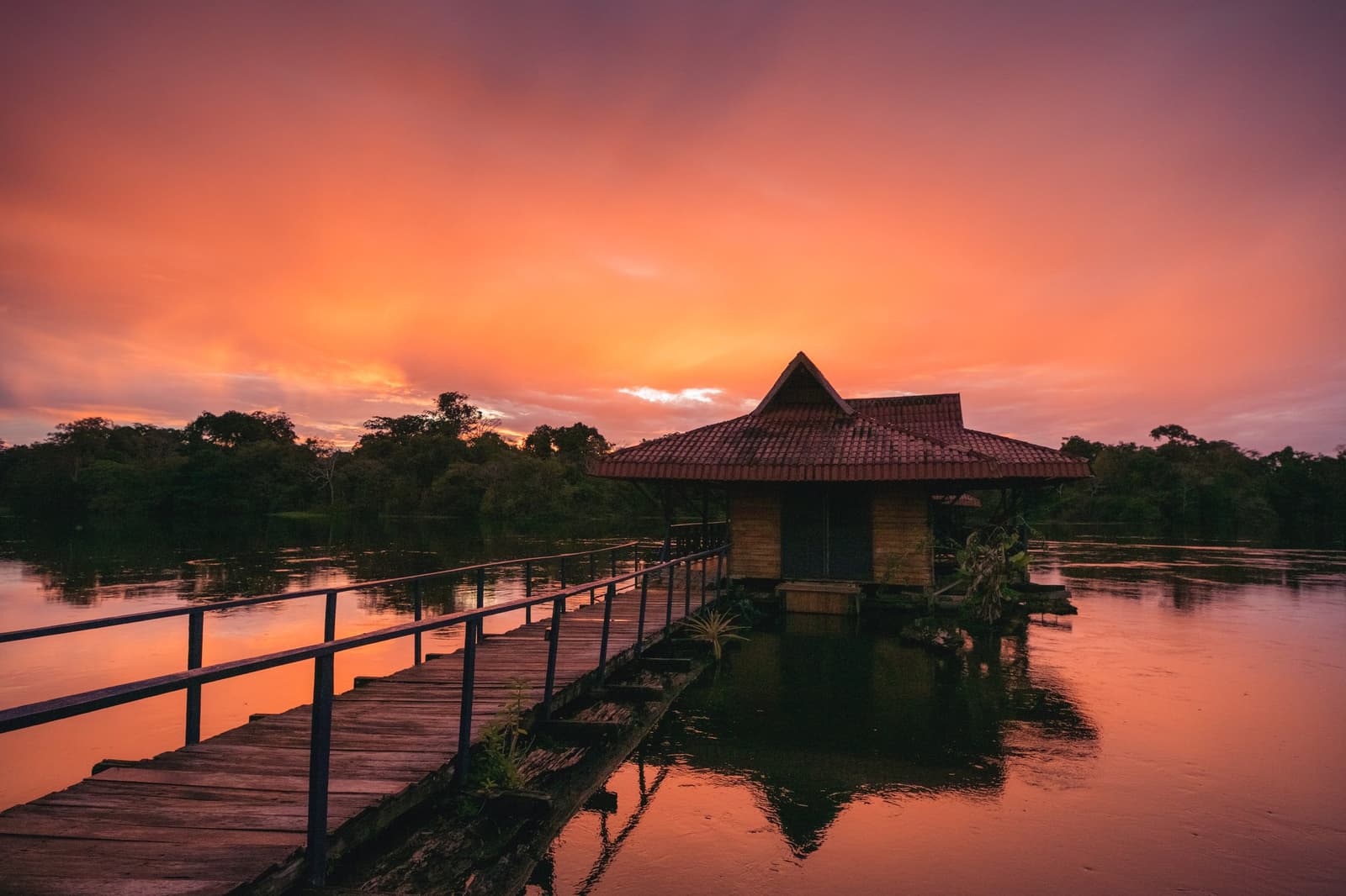 Your last thought before dinner, every single night. Photo: Guilherme Gomes A wooden boardwalk leads to an overwater bungalow with a terracotta-tiled roof on a calm jungle lake during an amazon rainforest ecolodge trip in brazil, the sky blazing orange and pink at sunset with a dense tree line reflected in the still water