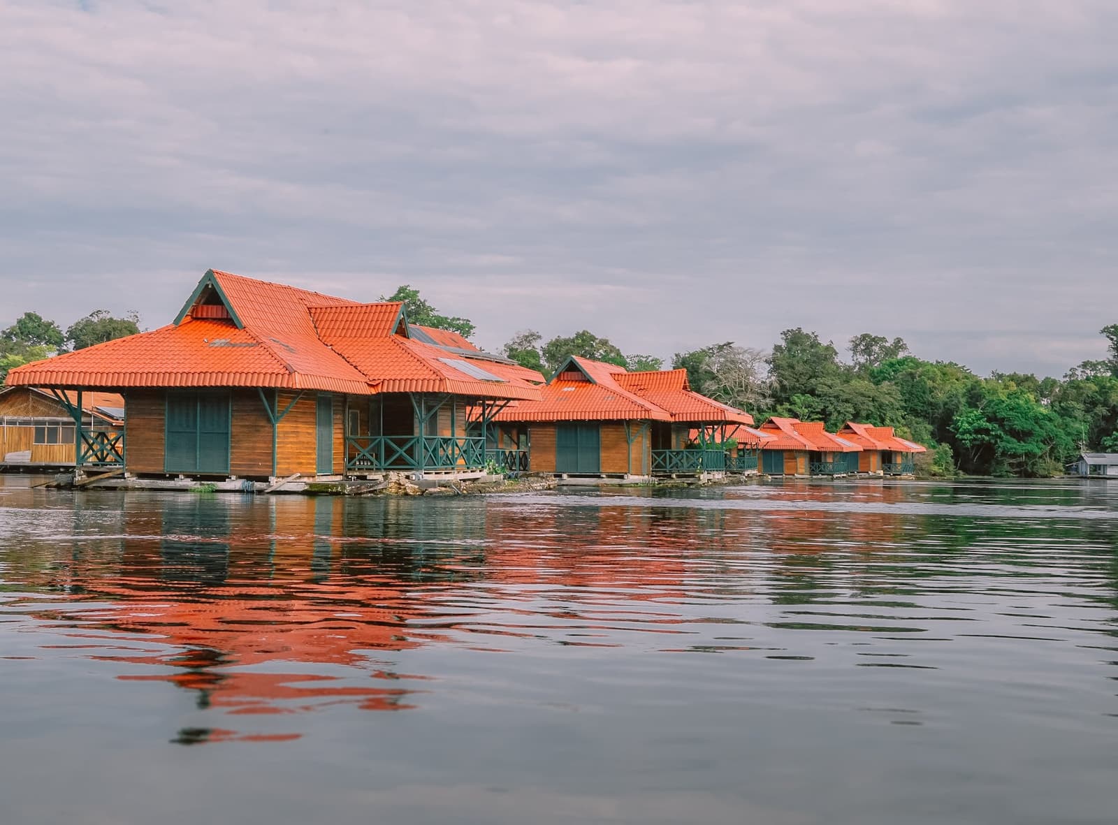 Red roofs on the water, forest at the door. Home for the week. A row of wooden cabins with terracotta-tiled roofs sitting on the water's surface at the Uakari floating lodge, their orange reflections rippling across a calm river with dense forest behind