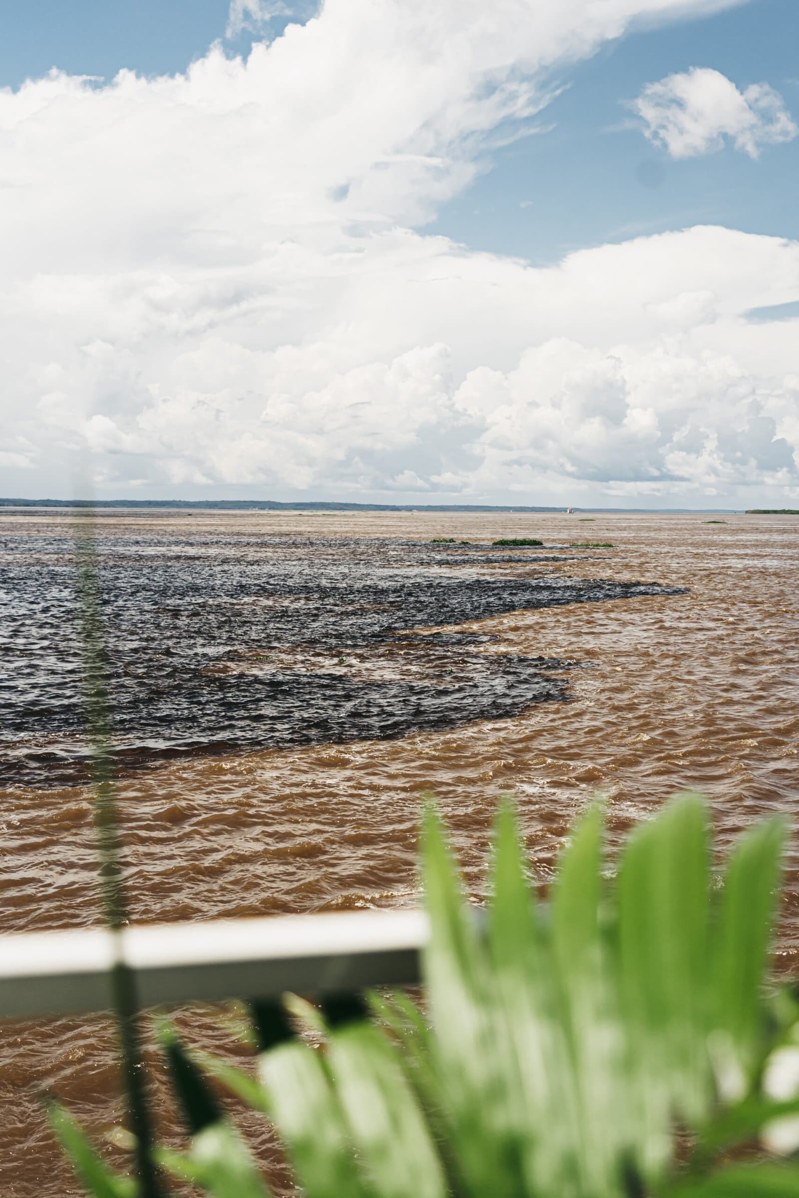 Two rivers, one surface, and neither one budges. The Meeting of the Waters seen from a boat rail, where the dark black Rio Negro and the sandy-brown Solimões flow side by side without mixing under a wide cloudy sky on a luxury Amazon cruise in Brazil