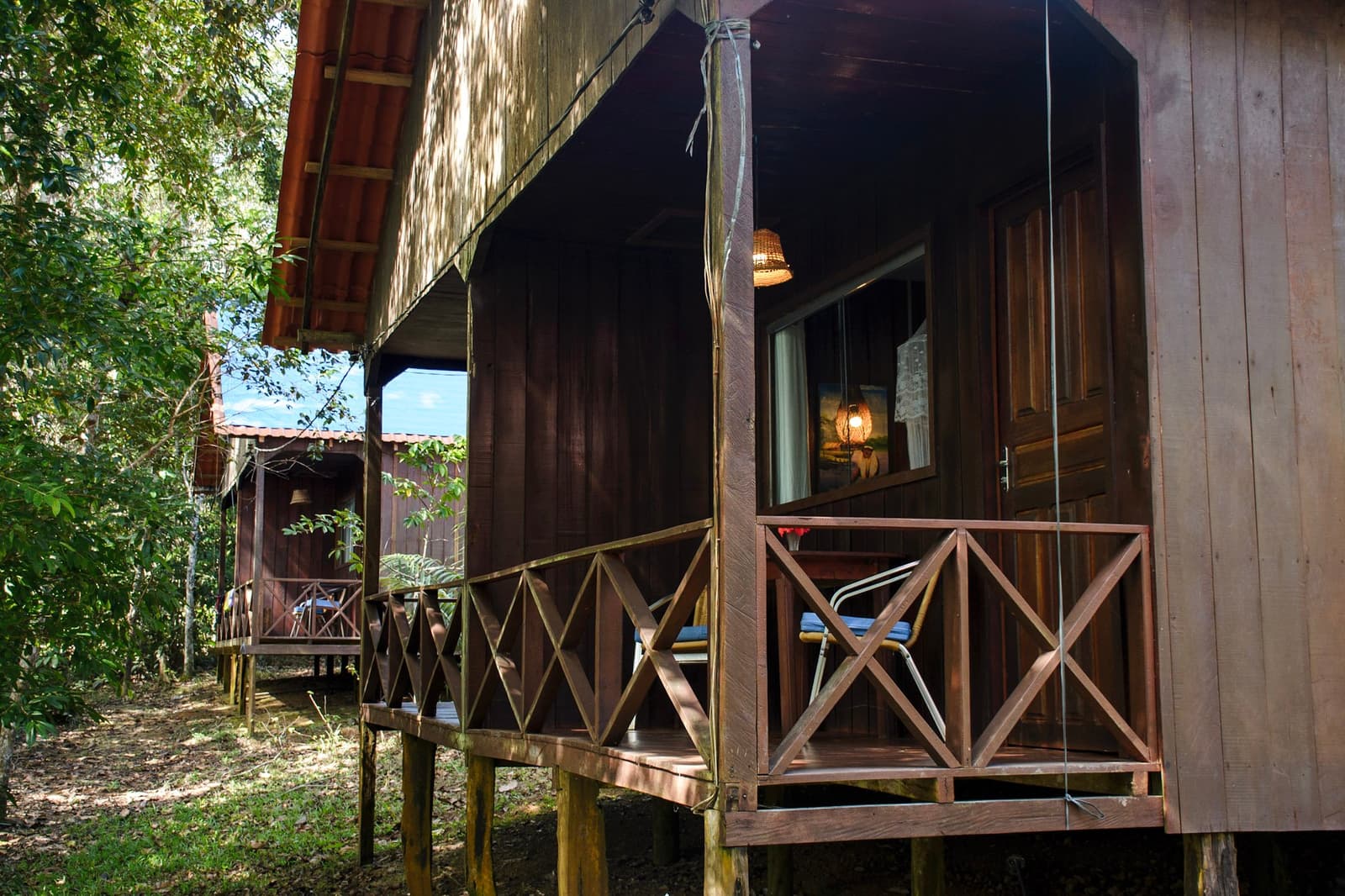 Simple, solid, and tucked into the trees, your base before the wild really begins. Two raised wooden cabin bungalows with cross-railed verandas, nestled side by side in dappled forest shade on this amazon and pantanal tour, warm lamp light visible through the window of the nearest unit