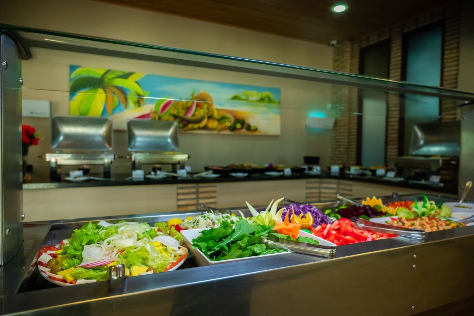 Fuel for long days on the water, served fresh and without compromise. A well-stocked salad buffet counter on an amazon and pantanal tour, with trays of fresh lettuce, spinach, red cabbage, sliced tomatoes, and carved vegetable garnishes under a glass sneeze guard, warm dishes visible in the background