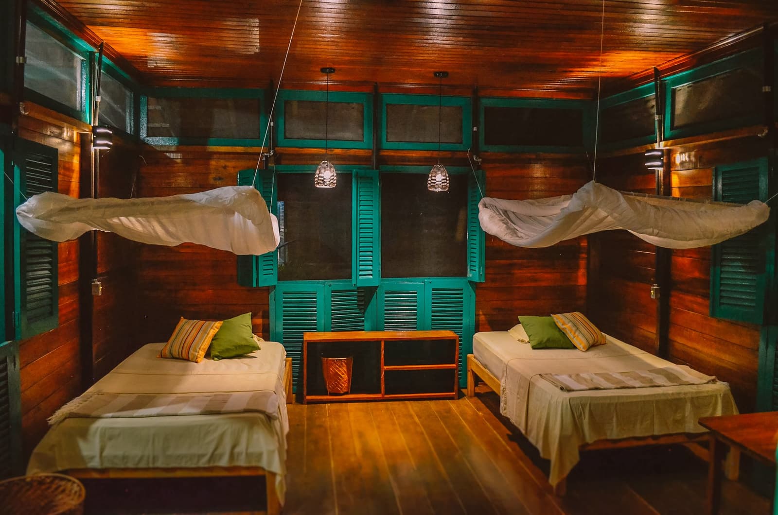 Rustic comfort done right: wood, net, and a good night's sleep in the forest. Twin-bed room at the Uakari Lodge featuring warm hardwood walls and ceiling, teal louvred shutters, mosquito nets suspended above each bed, and woven pendant lights casting a soft amber glow