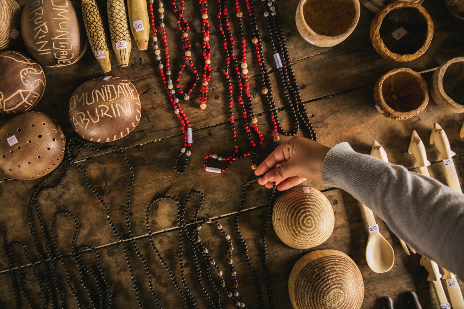Every piece on this table goes directly back to the family who made it. Photo: Guilherme Gomes Overhead view of a wooden table covered with handmade crafts from Comunidade Caburini, including red and dark seed-bead necklaces, carved gourd rattles, woven palm baskets, wooden spoons, and bone figurines, as a visitor's hand reaches in to browse during an amazon rainforest ecolodge trip in brazil