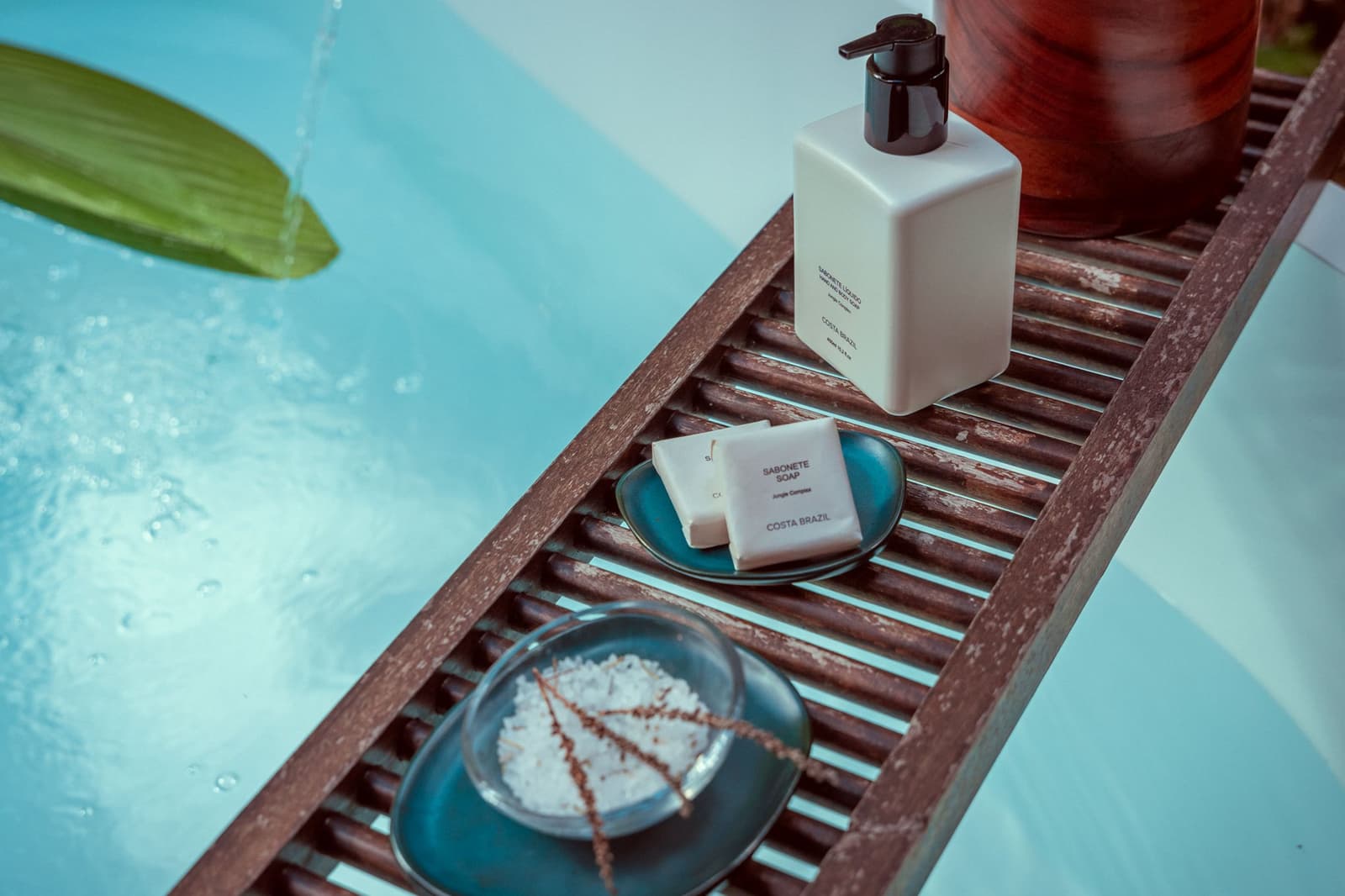 Costa Brazil in the jungle. Someone at this lodge really thought about your shower. Photo: Samuel Melim A wooden bath tray resting beside a turquoise pool holds Costa Brazil Jungle Complex soap bars on a teal ceramic dish, a white pump-dispenser bottle, and a small plate of bath salts with a dried botanical sprig, with a tropical leaf floating in the water alongside — thoughtful amenities on a luxury Pantanal and Amazon trip
