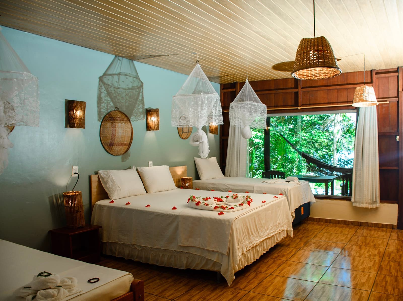 Flower petals on the bed, a hammock just outside the window. The forest can wait five minutes. Comfortable jungle lodge room on an amazon and pantanal tour, featuring two white beds decorated with red flower petals and towel art, lace mosquito nets hanging from the ceiling, wicker pendant lights, handwoven wall décor, and a large window overlooking a hammock on a green forest balcony