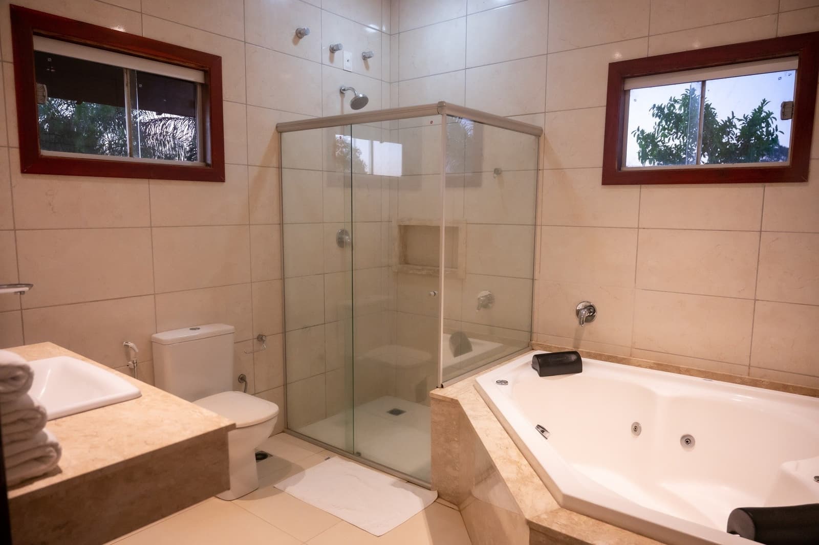 Hot shower or jacuzzi after a day on the river, your call. Spacious en-suite bathroom at a Pantanal lodge on an amazon and pantanal tour, featuring a glass-enclosed shower, corner jacuzzi tub with marble surround, vanity with folded towels, and two windows looking out to greenery