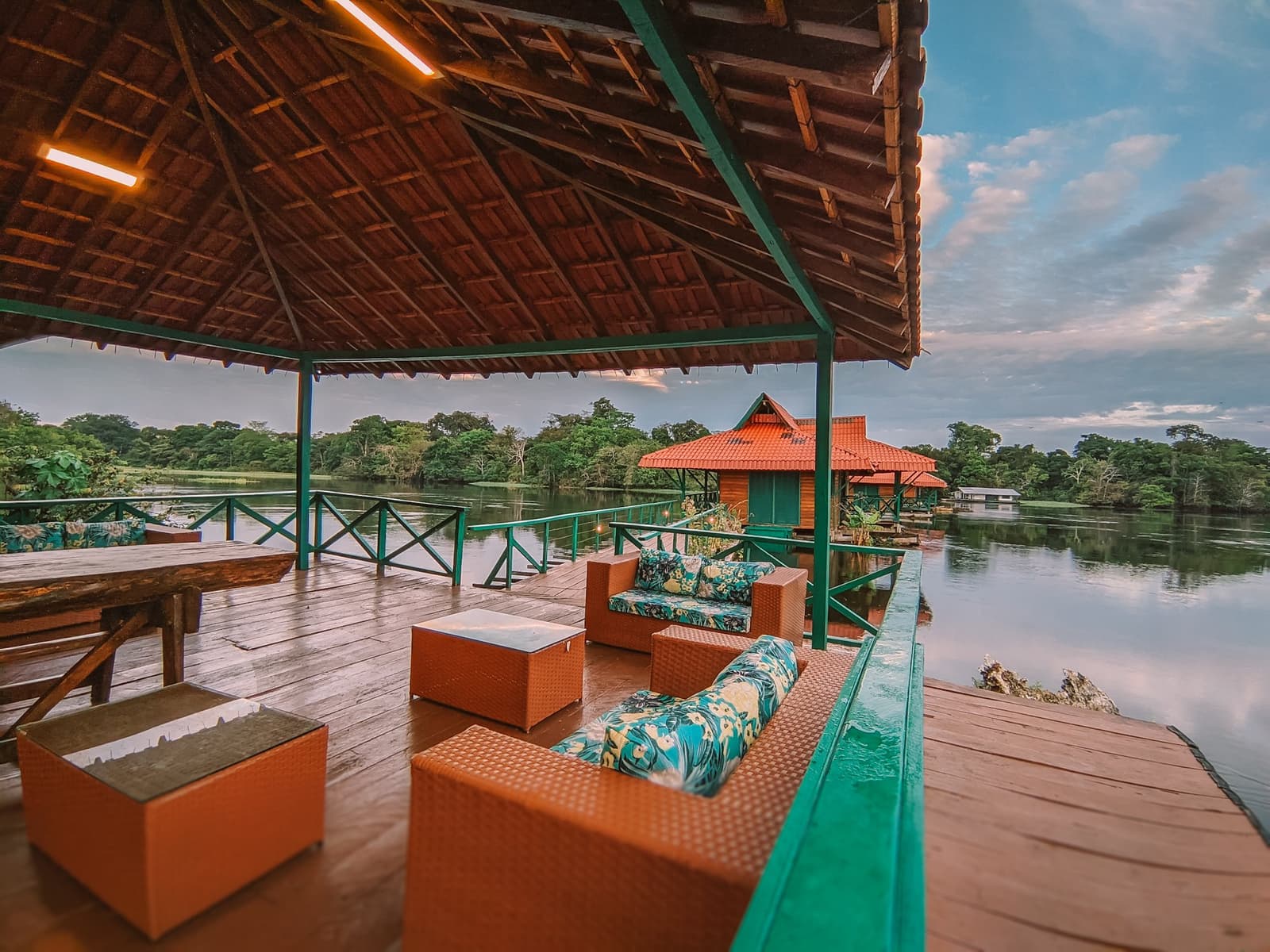 Sundowners hit different when the Amazon is your garden fence. Covered wooden deck of the Uakari floating lodge on an amazon rainforest ecolodge trip in Brazil, with rattan sofas and floral cushions overlooking a calm river channel and forested banks at dusk