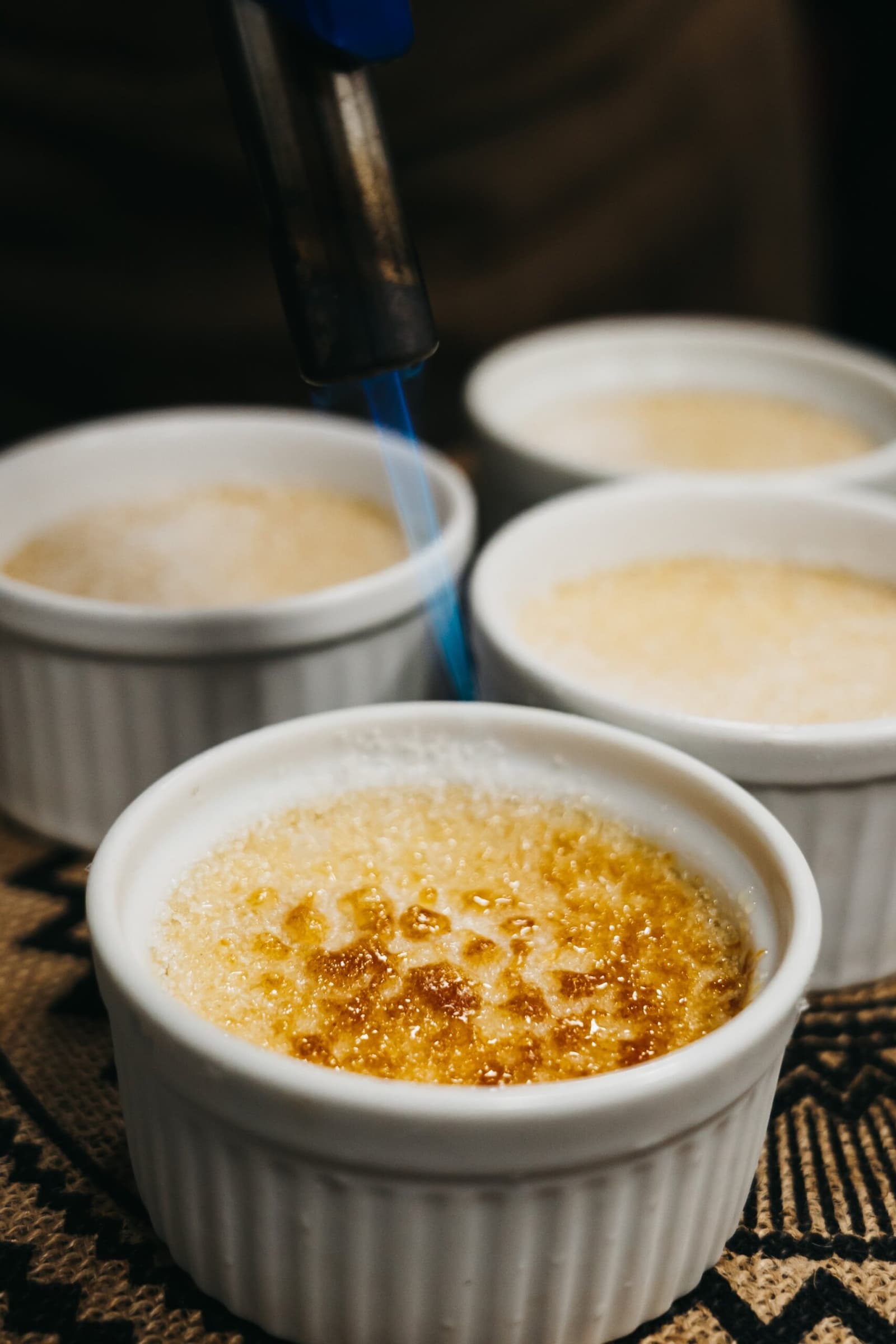 Gourmet dessert, river in every direction, no complaints. A kitchen torch caramelizing the sugar topping on a crème brûlée ramekin, with three more unfinished ramekins waiting in the background, served on a patterned textile aboard a luxury Amazon cruise in Brazil.
