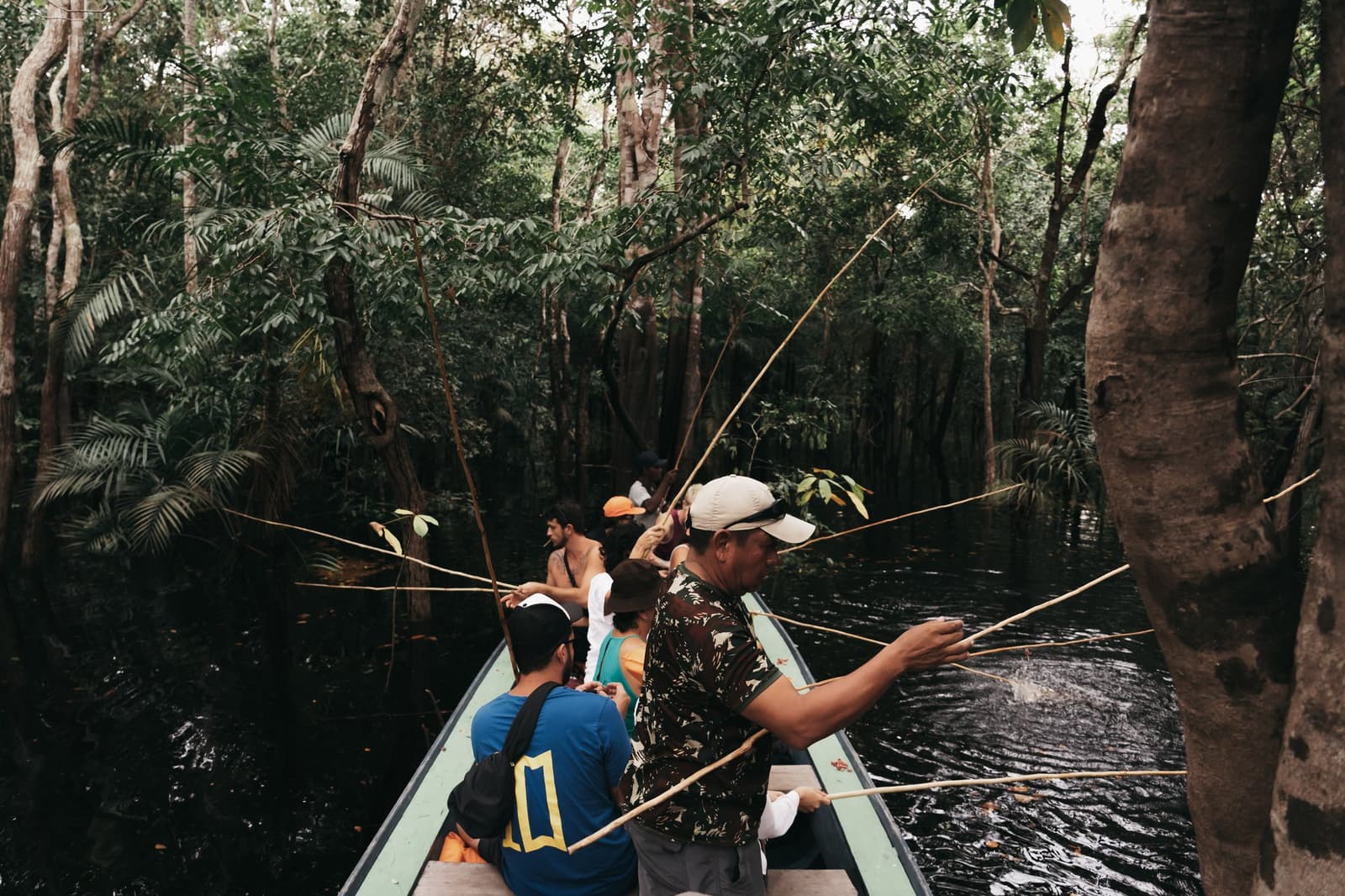 Everyone's line in the water, nobody's caught anything yet. Perfectly fine. A group of travelers fishing with bamboo rods from a narrow canoe on a dark blackwater channel, surrounded by dense flooded forest on a luxury Amazon cruise in Brazil.