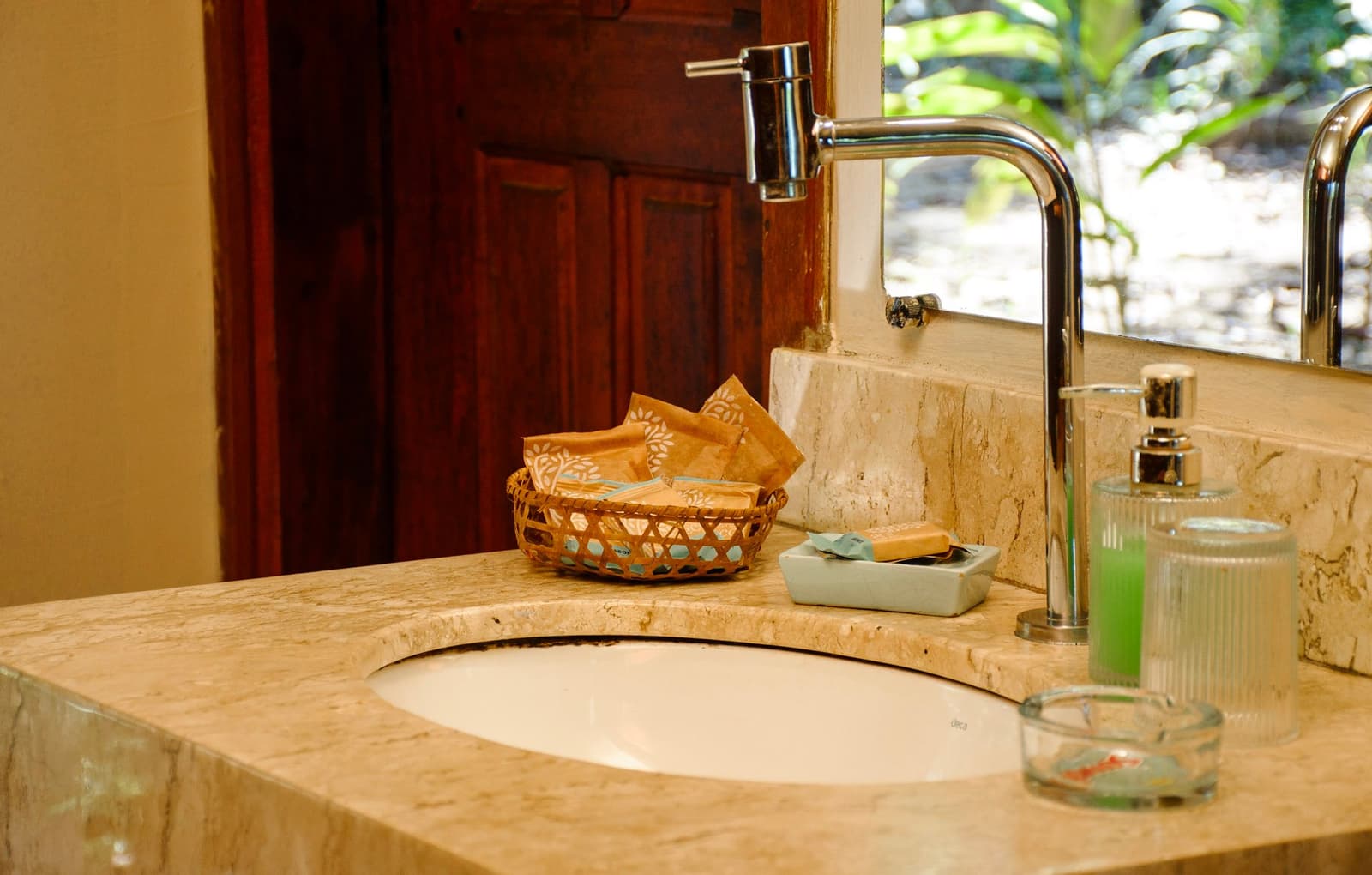 The little wicker basket of toiletries is a surprisingly nice touch after a long day outside. Stone-topped bathroom vanity at a lodge on an amazon and pantanal tour, with a chrome faucet, wicker amenity basket, and green toiletries beside a window overlooking tropical foliage