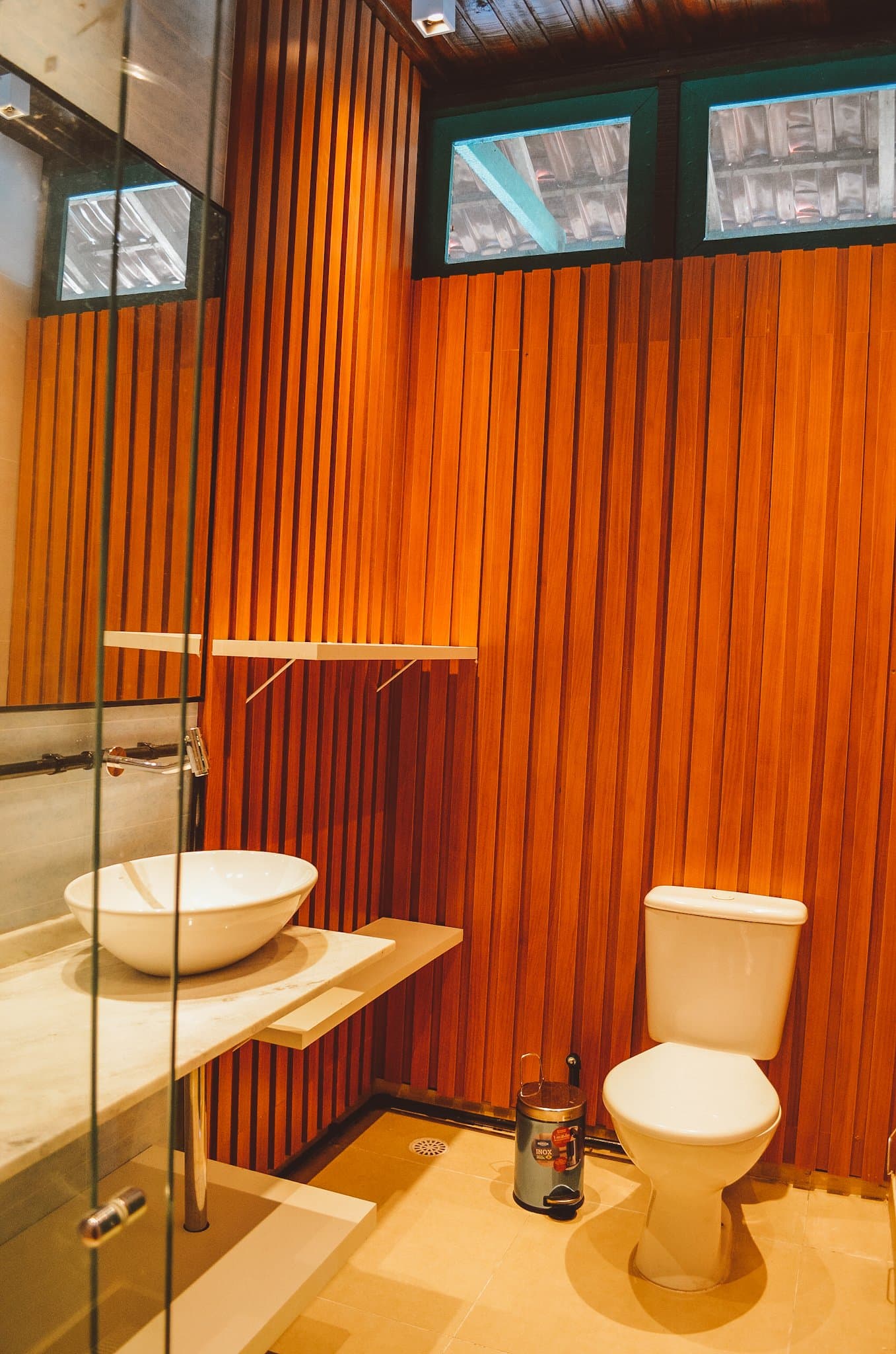 Warm timber walls, a proper sink, and enough light to find your sunscreen. Private bathroom at the Uakari Lodge during an amazon rainforest ecolodge trip in brazil, featuring warm timber slat walls, a white vessel sink on a marble countertop, glass shower partition, and glass-block transom windows
