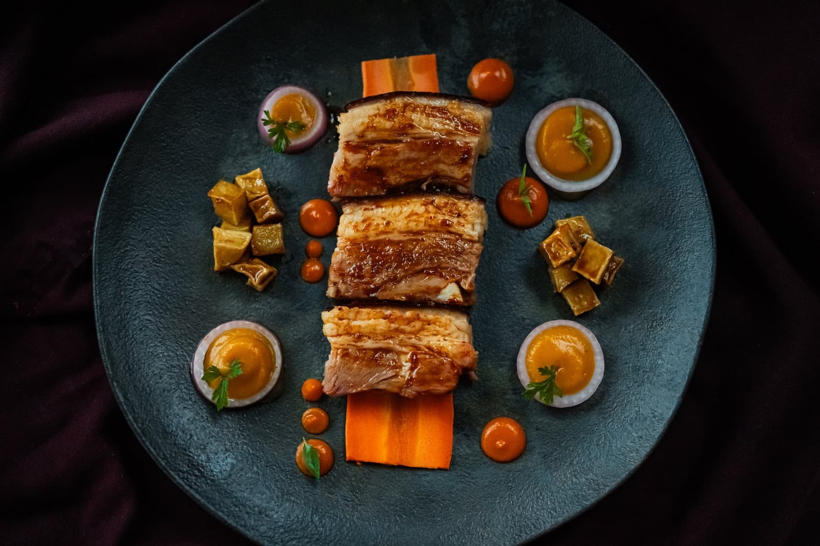 Proof that eating well at sea has nothing to do with proximity to a city. Three glazed pork belly slices plated on a teal dish with carrot ribbons, golden sauce cups, cherry tomatoes, and braised vegetable cubes, served on the luxury Amazon cruise in Brazil