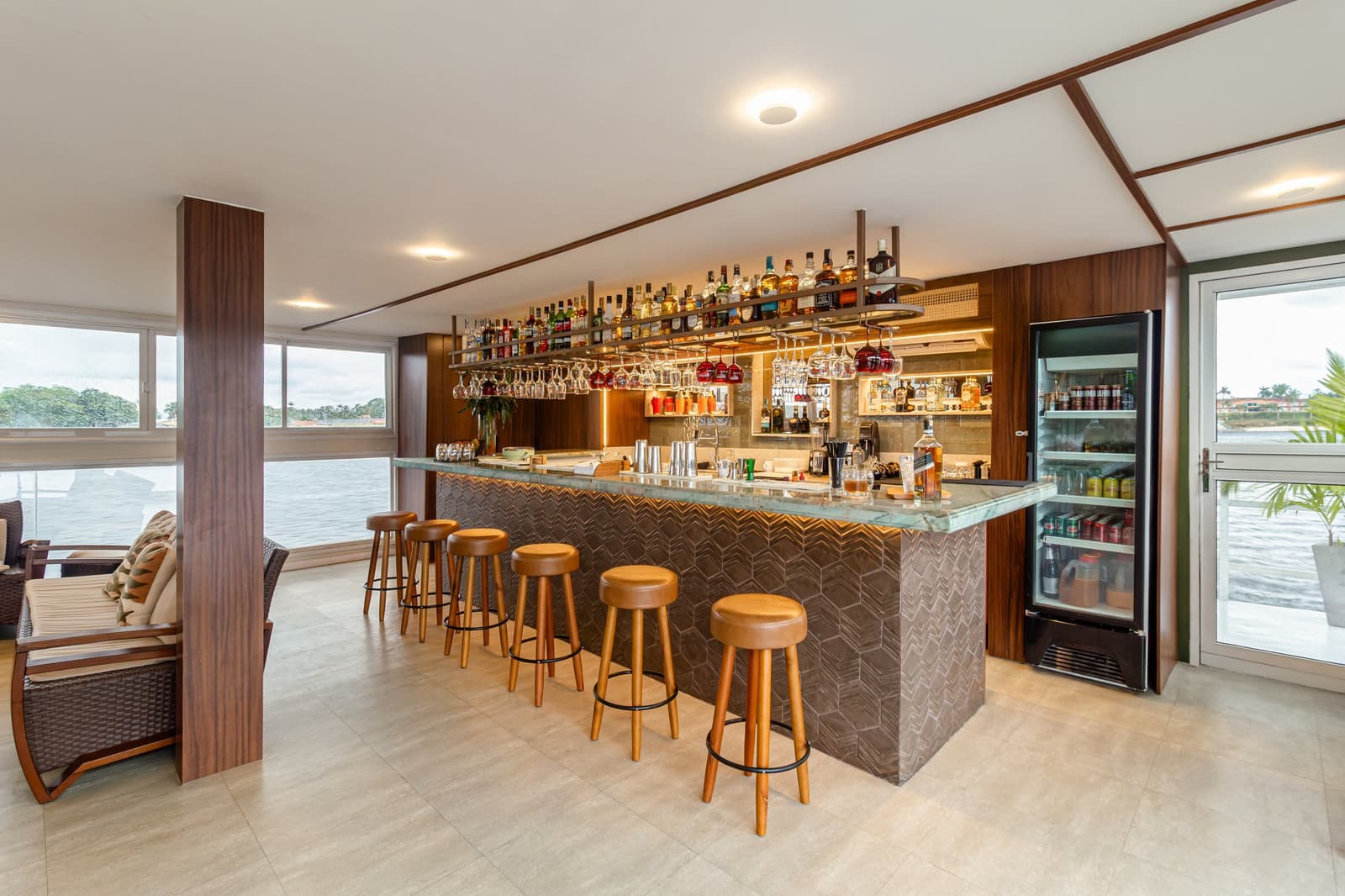 A proper bar, stocked and ready, with the river right outside the window. Photo: STUDIO CREATE Well-stocked bar on a luxury Amazon cruise in Brazil, featuring a green marble counter, rows of spirit bottles on backlit shelves, hanging wine glasses, leather bar stools, and panoramic windows overlooking the river