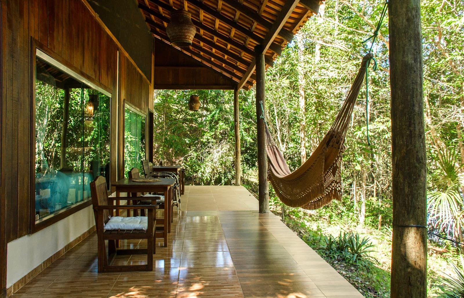 A hammock at eye level with the canopy. Hard to argue with that logic. Covered veranda of an amazon jungle lodge with a fringed cotton hammock hanging between wooden posts, a teak dining table and chairs, wicker pendant lights, and dense green forest just beyond the open edge