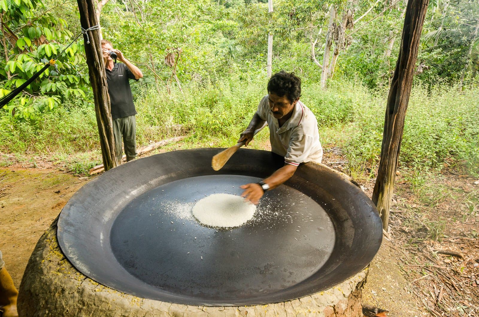 Farinha, the flour that feeds the Amazon, made right in front of you. Photo: Leonide Principe A man toasts cassava flour on a large iron griddle outdoors during an amazon rainforest ecolodge trip in brazil, while a visitor photographs the scene against a lush green forest backdrop.