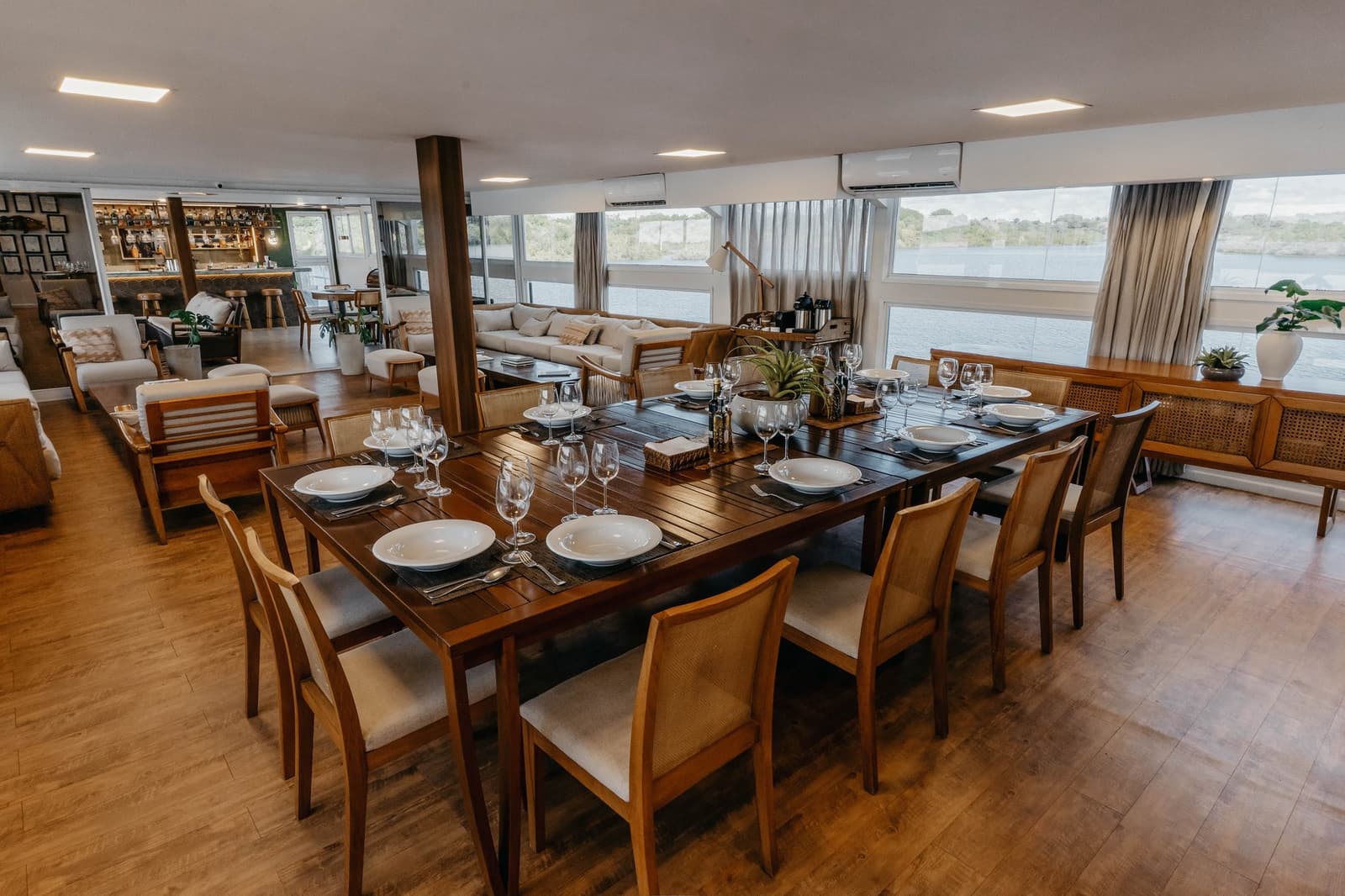 Lunch is served, and the Rio Negro is right outside the window. Formally set dining table with white porcelain and wine glasses aboard a luxury Amazon cruise in Brazil, with a lounge area and wide river-view windows beyond