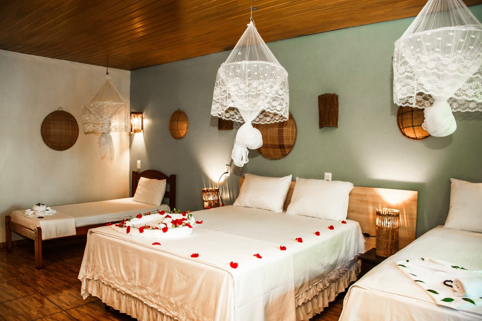 Rose petals and lace nets, someone put real thought into your welcome. A triple-occupancy lodge room on an amazon and pantanal tour, with three beds dressed in white linens scattered with red rose petals, lace mosquito nets hanging from a wood-panelled ceiling, wicker wall décor, and warm bamboo lanterns glowing on the nightstands.