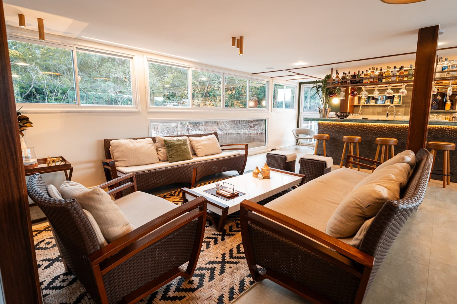 Sundowners, a proper bar, and the forest sliding past the windows. Comfortable wicker and wood lounge seating with cream cushions on a patterned rug aboard a luxury Amazon cruise in Brazil, with a fully stocked bar and panoramic windows overlooking the river and rainforest