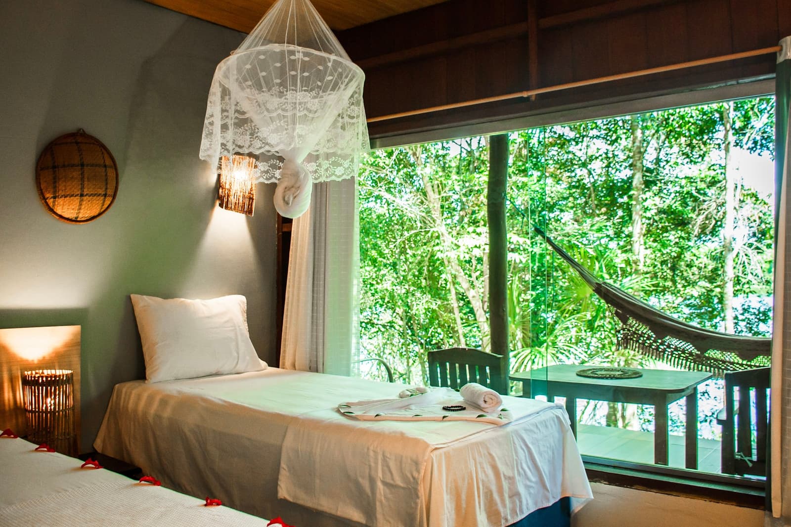 Lace nets, white linen, and a hammock waiting just outside the glass. Twin bedroom at an amazon jungle lodge with white linen beds under a lace mosquito net canopy, warm wall sconce, woven basket wall art, and a large window opening onto a green forest canopy with a hammock on the balcony