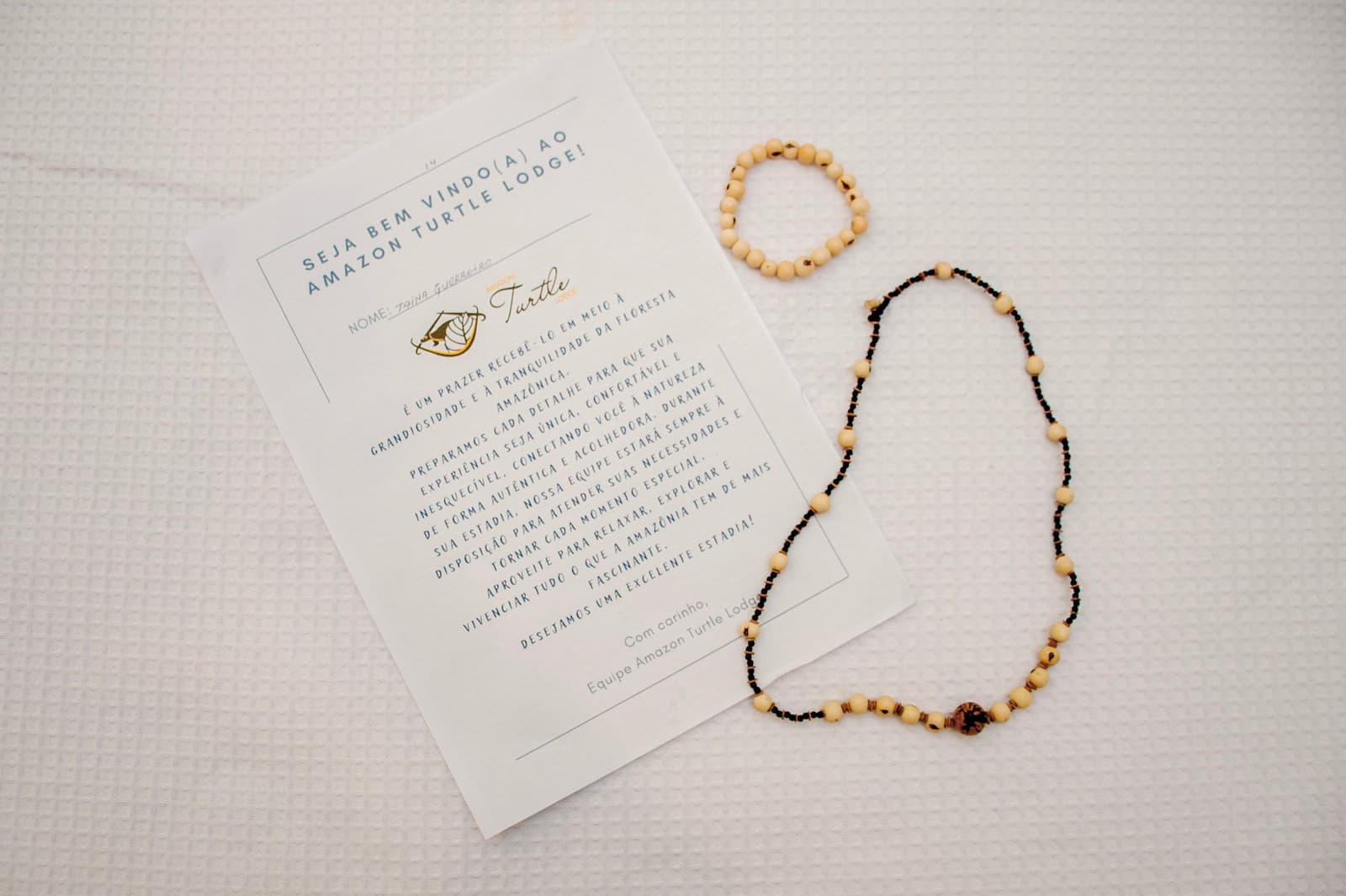 Handmade seeds, a note with your name on it. Someone was expecting you. Personalized welcome card from an Amazon jungle lodge beside a handmade seed bead necklace and matching bracelet on a white waffle-weave bedspread