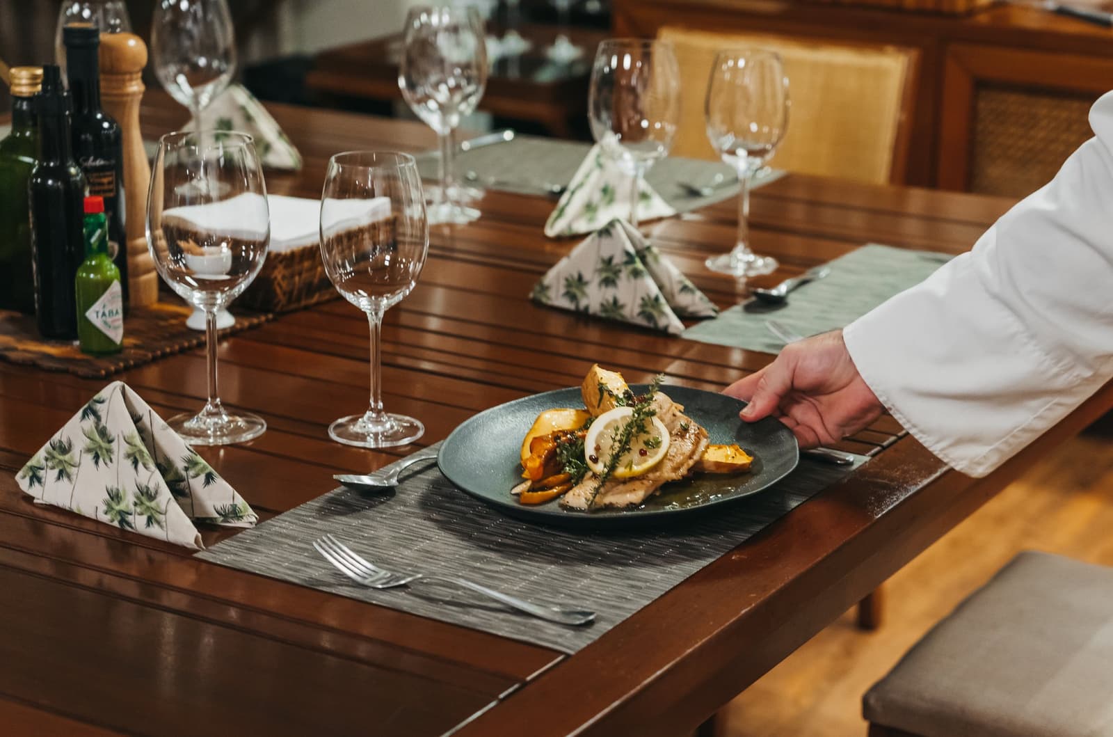 Gourmet plating, river outside, nothing left to wish for. A chef in a white jacket places a dark plate of grilled fish fillet with roasted orange peppers, lemon slices, and fresh herbs on a formally set wooden dining table aboard a luxury Amazon cruise in Brazil, with folded botanical-print napkins and wine glasses arranged for several guests