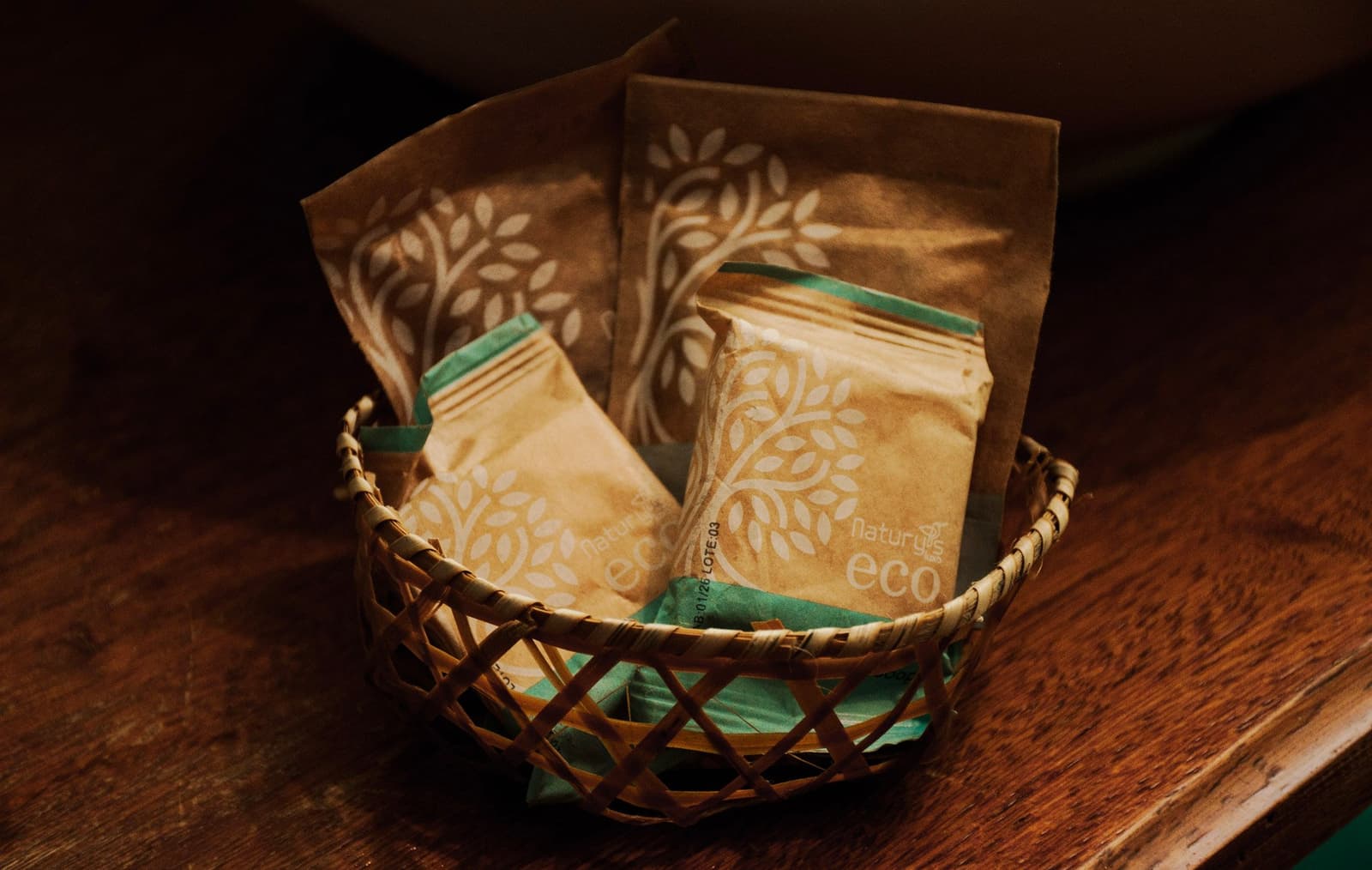 Plastic-free, thoughtfully packed. Even the little extras respect the forest outside. Four kraft-paper Natury Eco toiletry sachets with a leaf-print design arranged in a small woven basket on a wooden surface, provided as eco-friendly amenities on an amazon and pantanal tour lodge stay