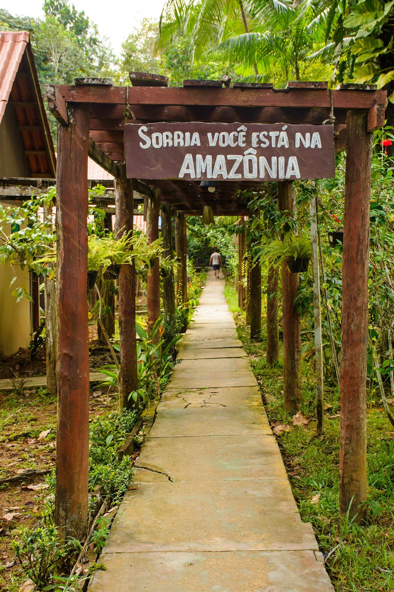 The forest has a sense of humor. Smile, you're in the Amazon. A rustic wooden pergola at an Amazon jungle lodge entrance displays a sign reading 'Sorria você está na Amazônia' — Smile, you are in the Amazon — with a tree-lined concrete path leading into the rainforest and a lone guest walking ahead on this amazon and pantanal tour.