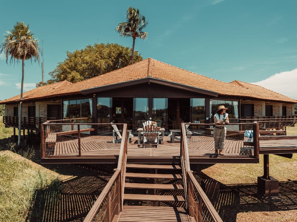 Photo: Felipe Castellari The main building of a Pantanal wildlife lodge with a tiled roof and a wide wooden deck under a bright blue sky, a guest standing between palm trees.