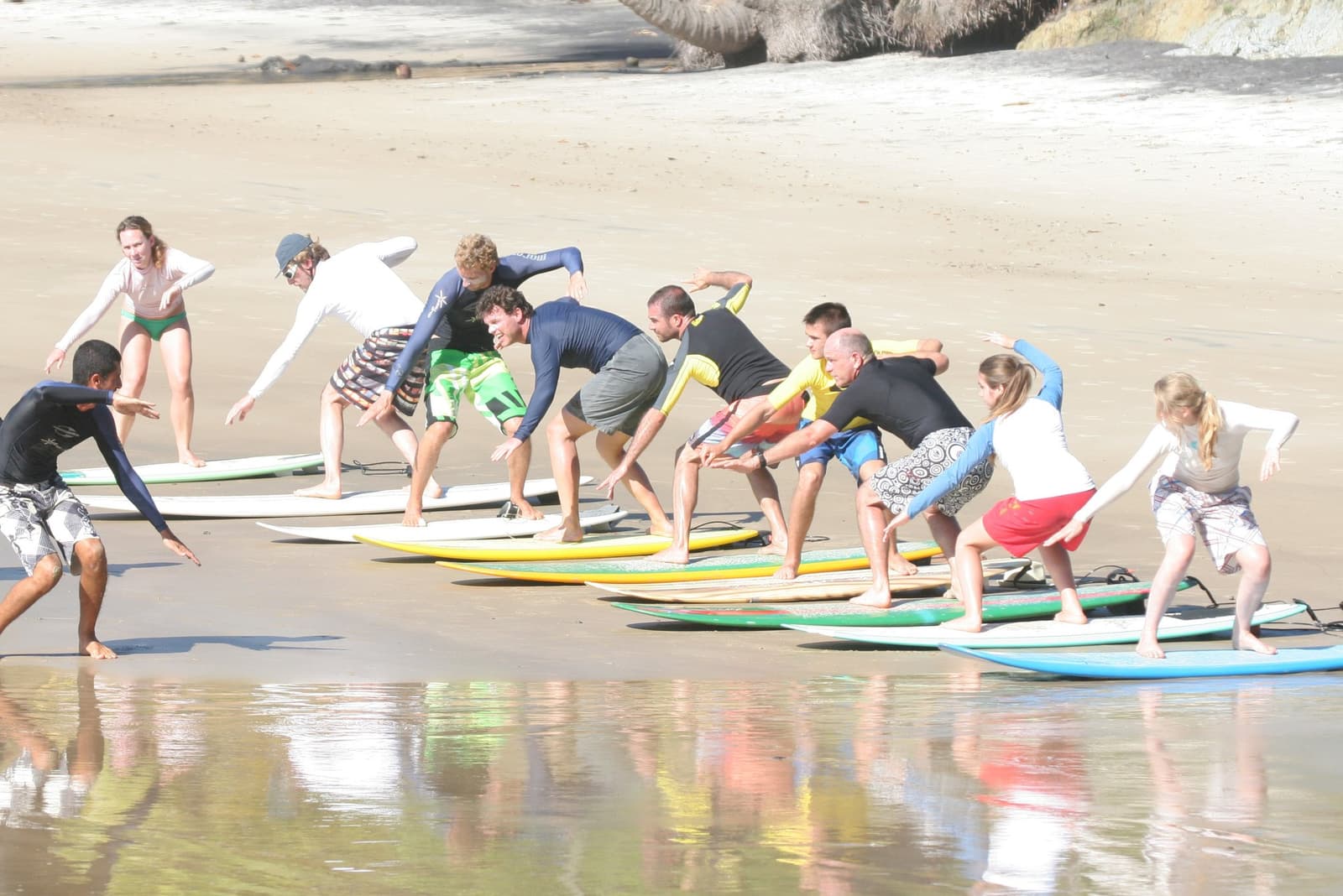 Everyone wobbles on the first try. That's the whole point. Photo: Picasa A group of about ten students practicing pop-up stance on surfboards during an itacaré surf school dryland drill on a sandy beach, with a local instructor demonstrating the crouched surf stance at the front of the line