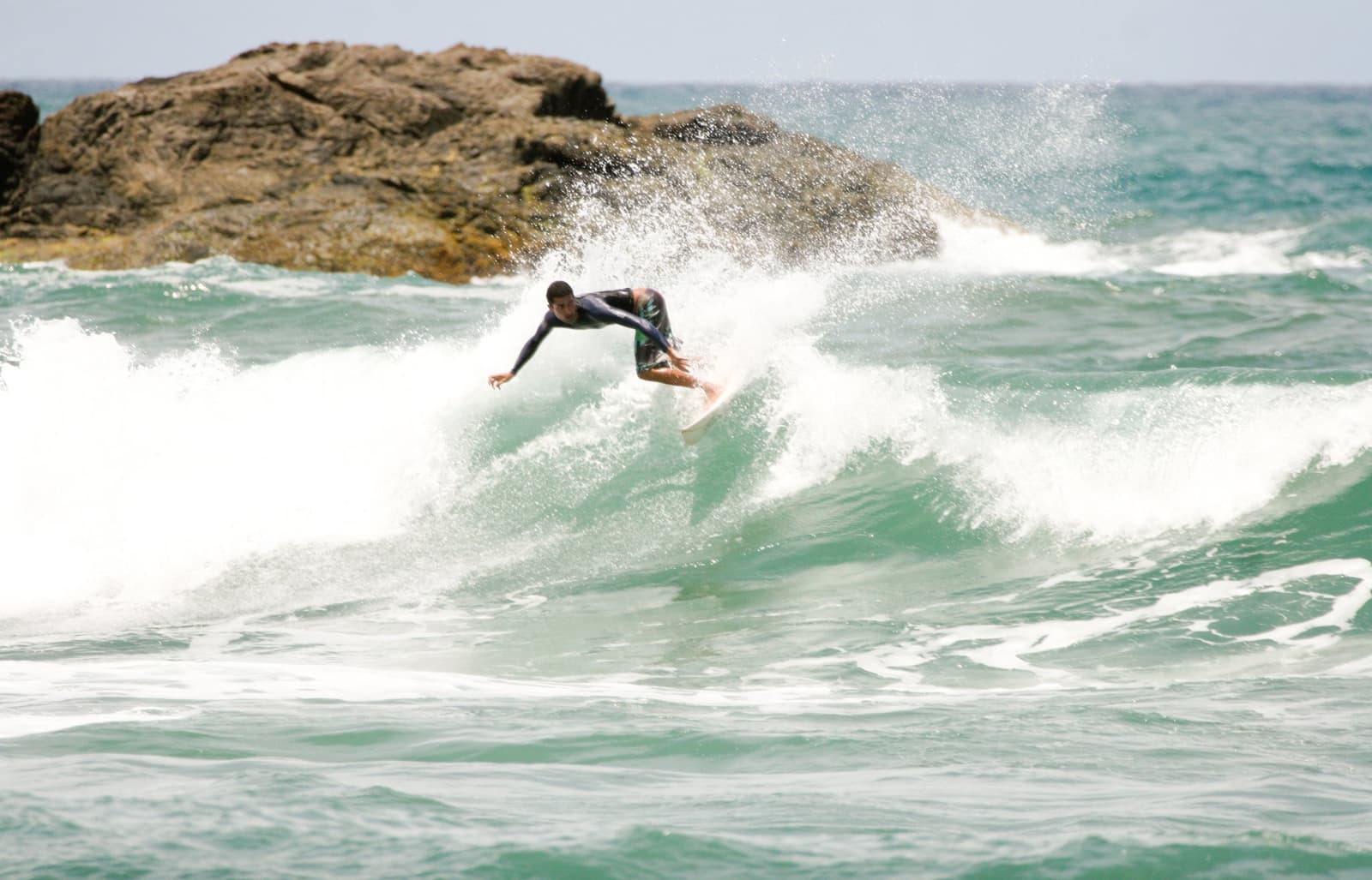 Outside the break, past the foam, this is what day three feels like. Photo: unknown A surfer in a long-sleeve rash guard executes a sharp cutback on a green wave at an itacaré surf school session, with a large rocky outcrop and white spray rising behind him