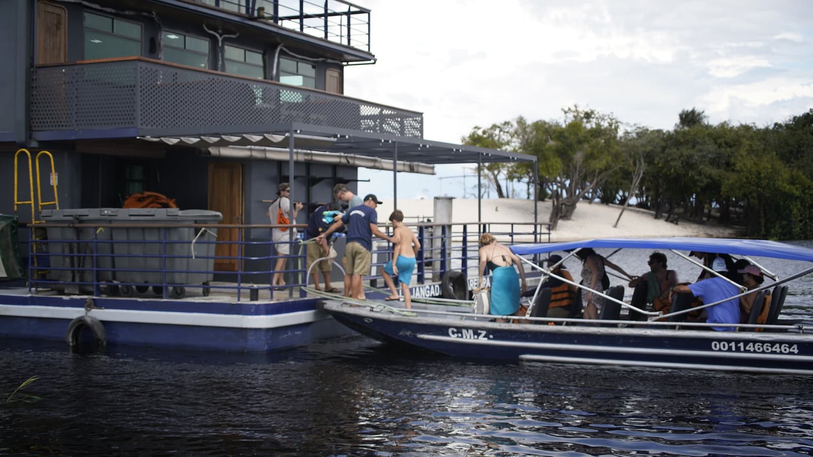 All aboard, and the Rio Negro takes it from here. Guests boarding a blue river speedboat at the La Jangada amazon cruise floating dock, with the two-storey steel catamaran and a sandy riverbank visible in the background