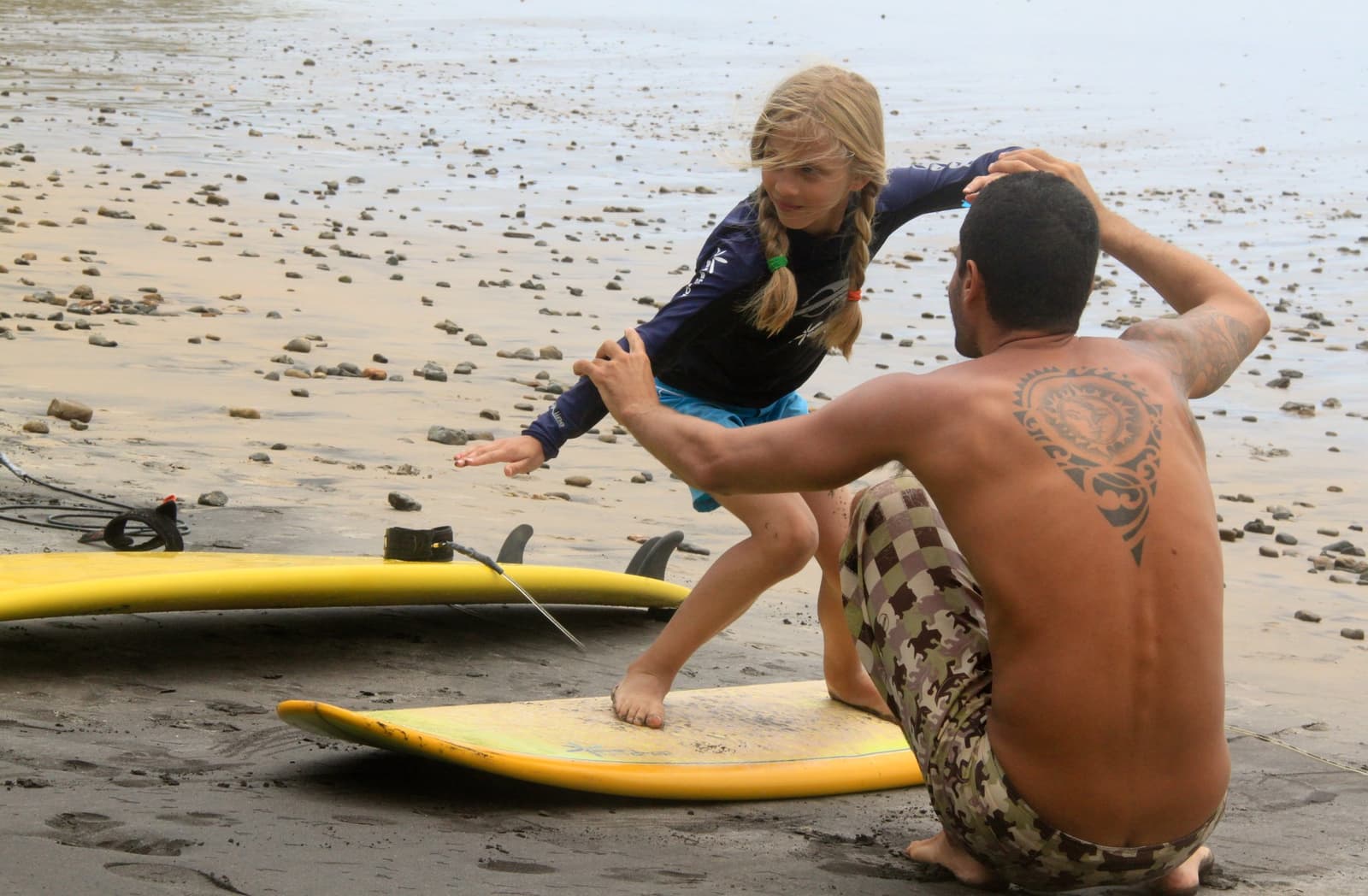 Getting the pop-up right on dry land first. Smart move. Photo: Picasa A surf instructor guides a young woman practicing her pop-up stance on a yellow surfboard on a dark sandy beach at the Itacaré surf school, with rocky tidal flats visible in the background.