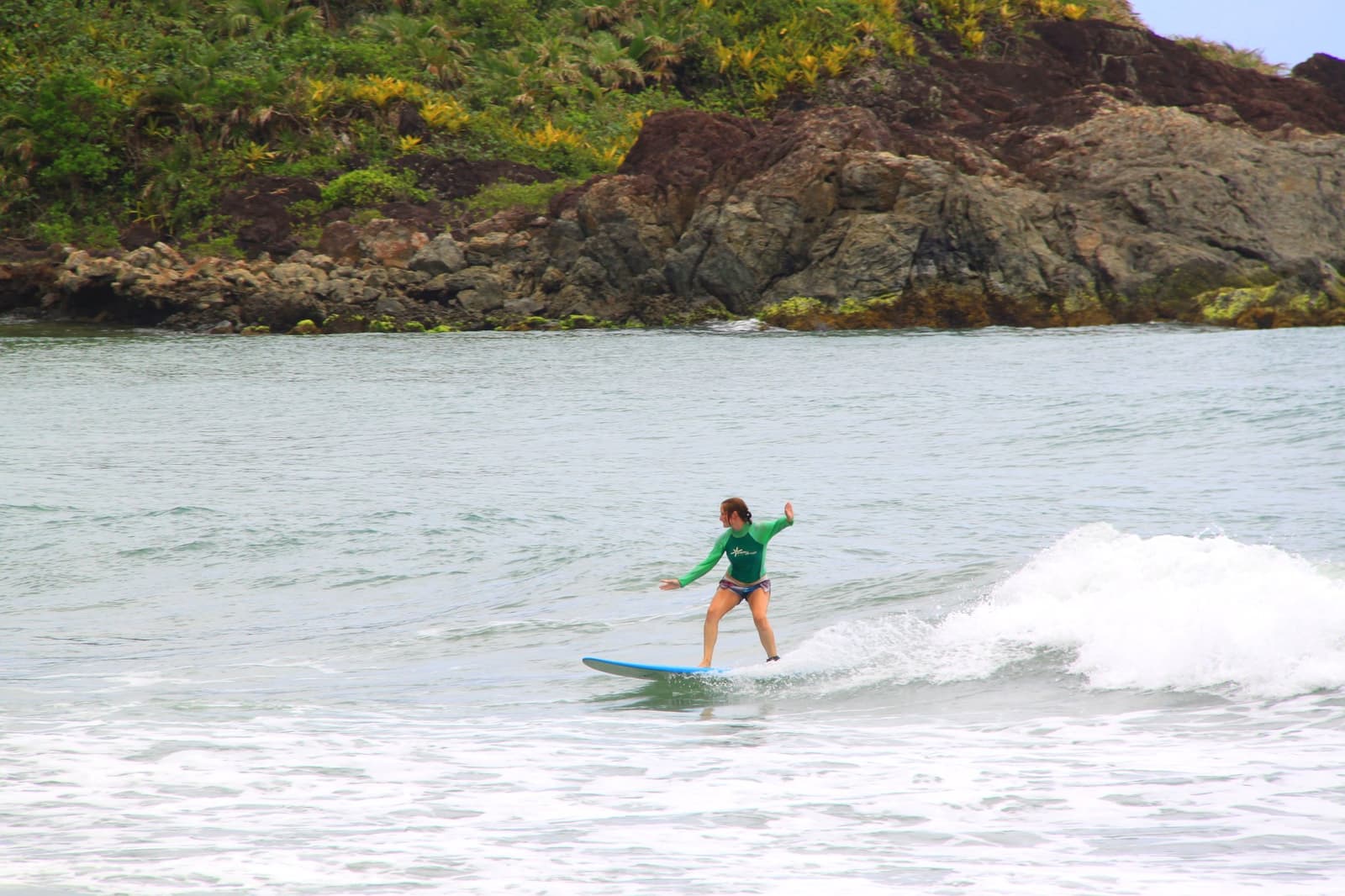 Arms out, weight forward, this is what clicking into place feels like. Photo: Picasa A woman in a green rash guard rides a small green wave on a blue longboard at an itacaré surf school session, with a rocky, vegetation-covered headland in the background.