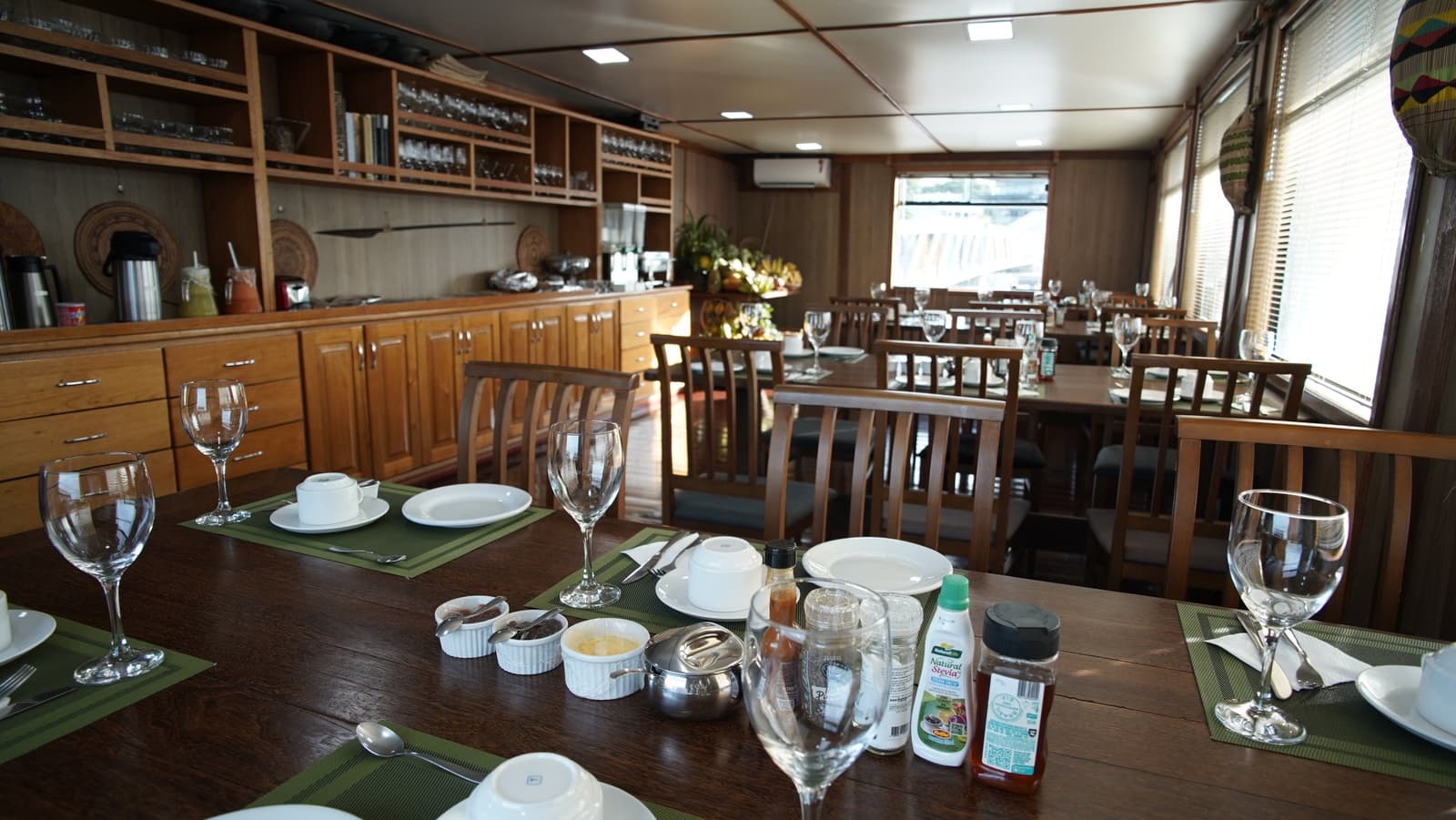 Good food, cold drinks, and the Rio Negro drifting past the window. Not a bad way to eat. The la jangada amazon cruise dining saloon set for a meal, with polished wood tables, green placemats, crystal wine glasses, white porcelain, condiments, and a full timber buffet along the wall