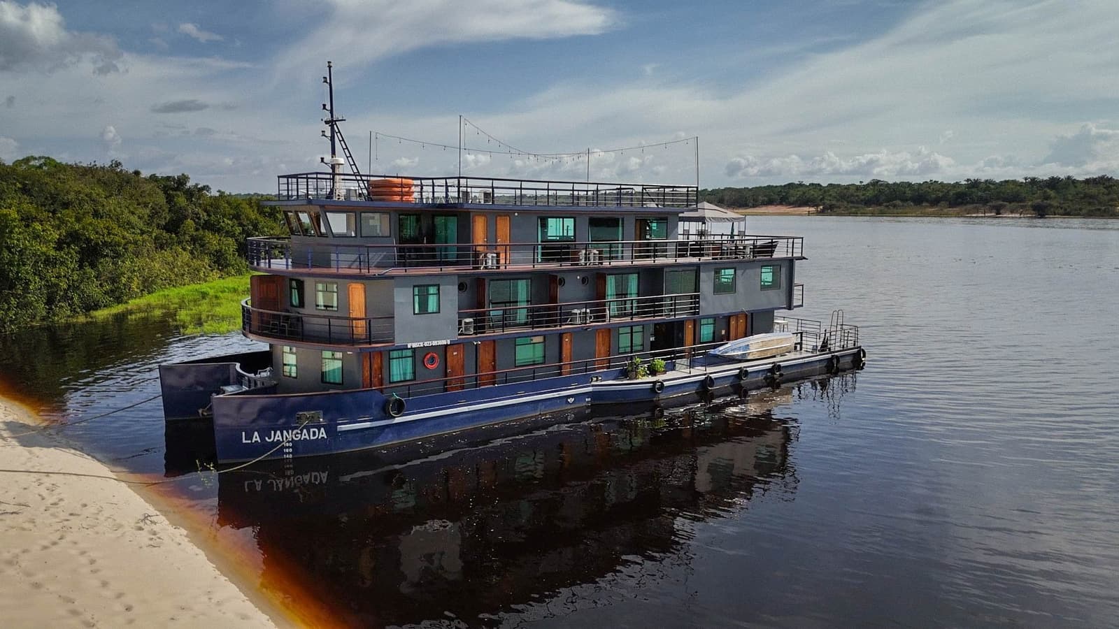 Three decks, twelve cabins, one very dark river, this is your ride. The La Jangada amazon cruise vessel moored on the dark waters of the Rio Negro, its three-deck steel hull and wood-trimmed cabins reflected in the river, dense rainforest lining the far bank under a partly cloudy sky