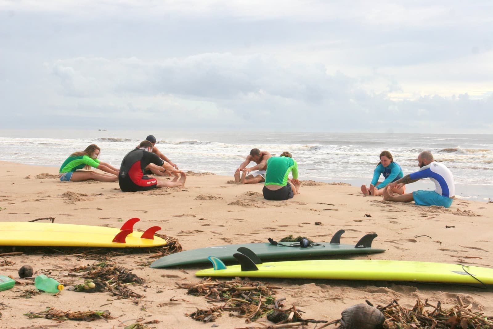 The warm-up nobody skips, your back will thank you later. Photo: Picasa Six students sitting on a sandy beach stretching in pairs before their itacaré surf school lesson, with surfboards laid on the sand in the foreground and waves breaking behind them