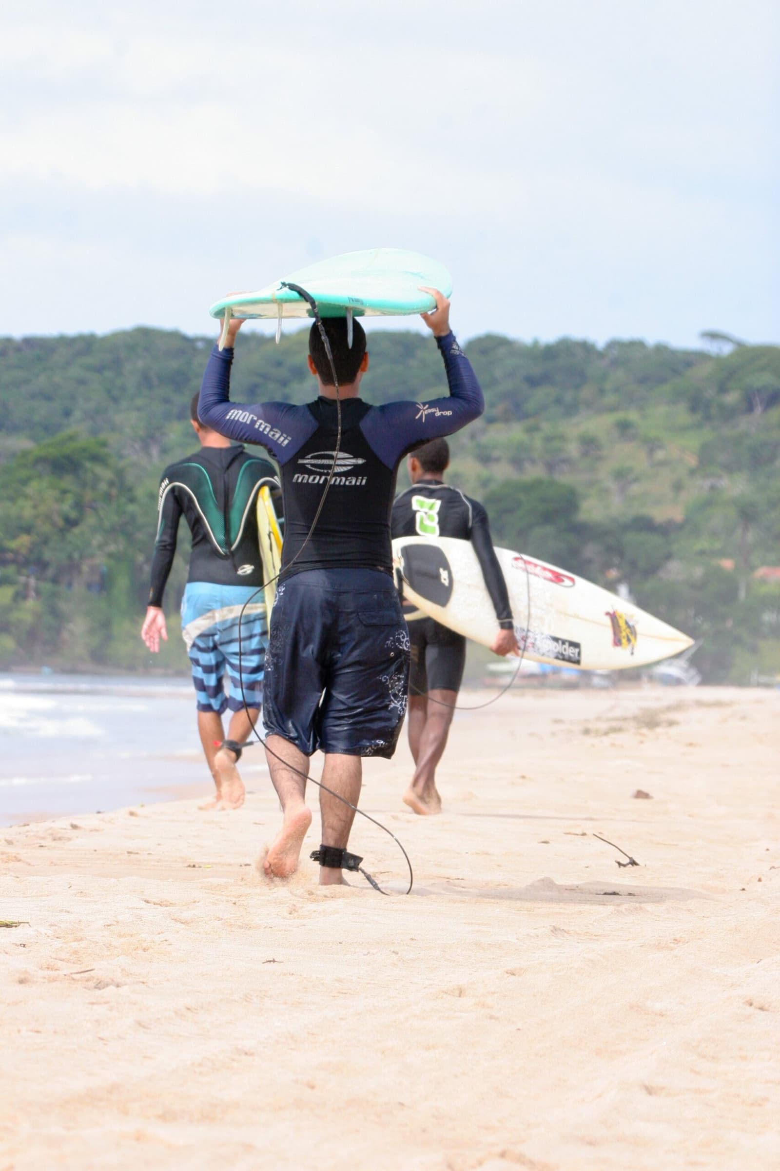 Boards up, leashes on, the lineup is calling. Photo: unknown Three surfers in wetsuits and boardshorts walking along a sandy beach toward the ocean at an itacaré surf school session, one carrying a turquoise board overhead, another holding a white shortboard, with dense green coastal forest in the background